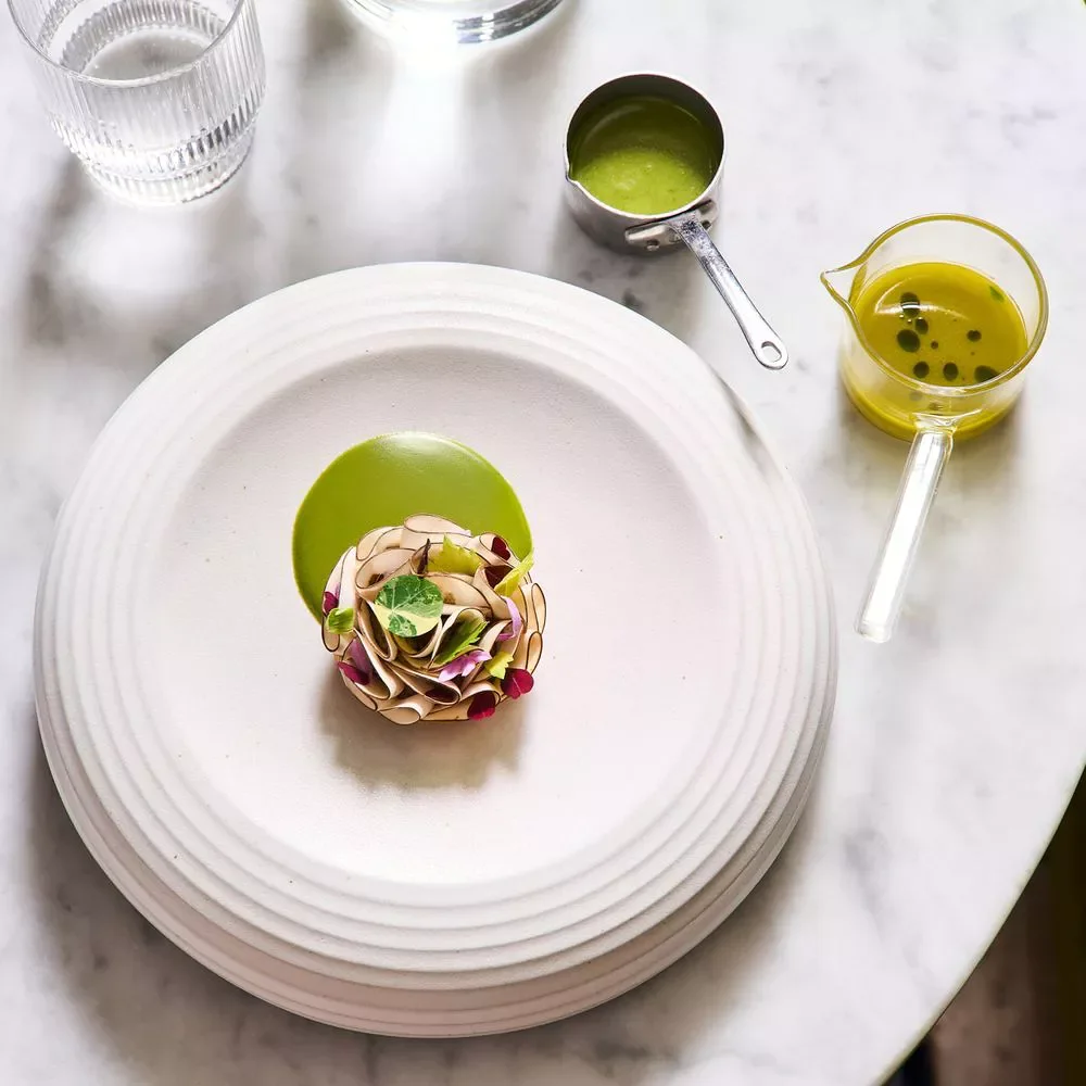 Une assiette blanche avec un plat élégant comprenant une fleur en spirale garnie de micro-pousses, accompagnée de deux sauces vertes à côté. Sur la table en marbre, il y a aussi des verres d'eau et des contains en verre avec de la sauce ou huile d'olive, une bouteille d'huile d'olive.