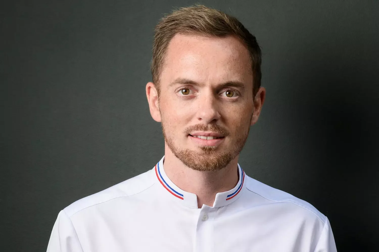 Portrait d'un homme jeune avec une barbe, portant une chemise blanche avec un col rayé rouge, blanc et bleu, devant un fond sombre.