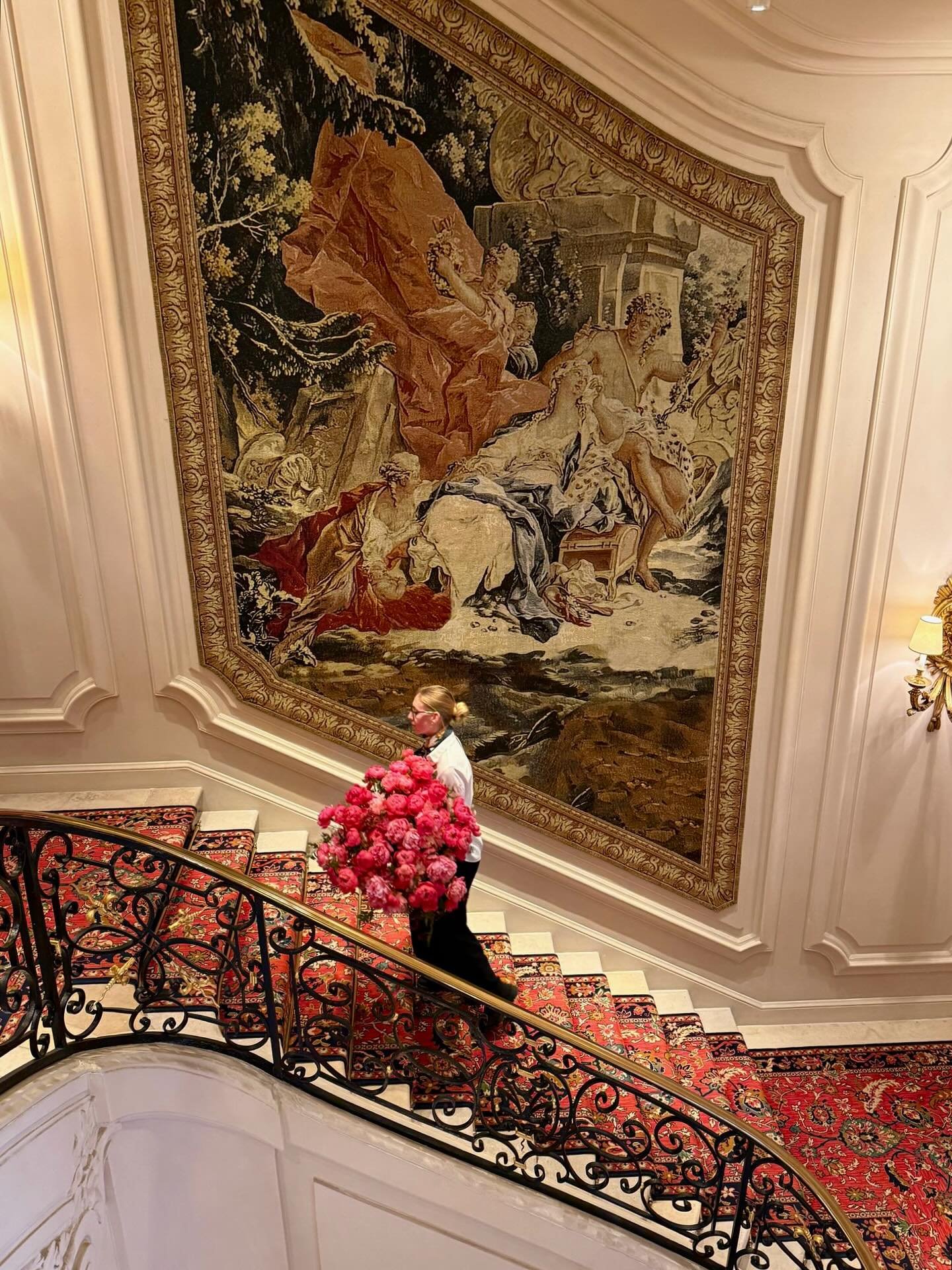 Une femme portant une grande gerbe de fleurs roses descend un escalier élégant décoré d'un tapis rouge et d'une rampe en fer forgé, avec un grand tapis mural représentant une scène historique ou mythologique en arrière-plan.