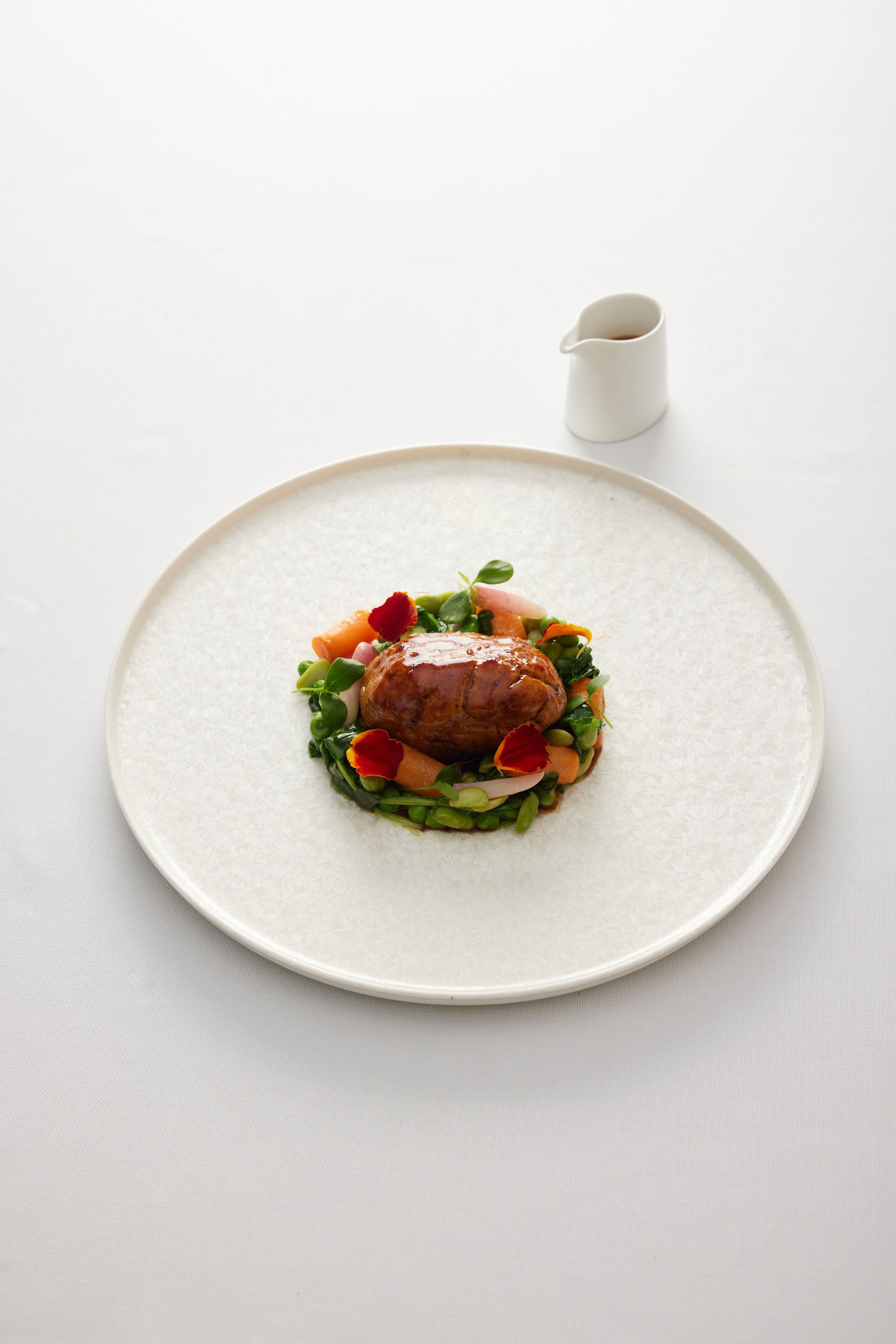 Plat gastronomique avec un morceau de viande, légumes et pétales de fleurs sur une assiette blanche élégante, garniture de sauce dans une cruche.