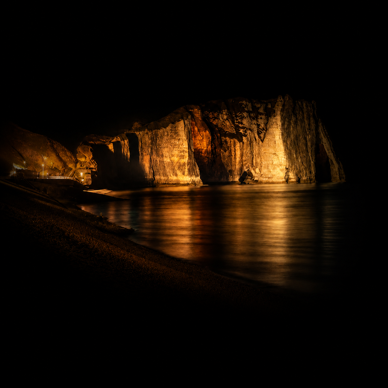 Falaises illuminées par des lumières dorées, reflet sur l'eau calme, scène nocturne côtière.