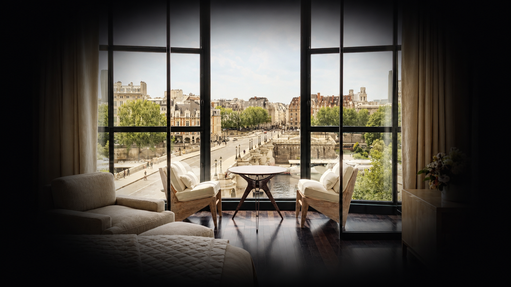 Une vue d'une ville depuis une fenêtre, avec une terrasse intérieure équipée de deux chaises rembourrées, une petite table, un canapé et des rideaux beiges.