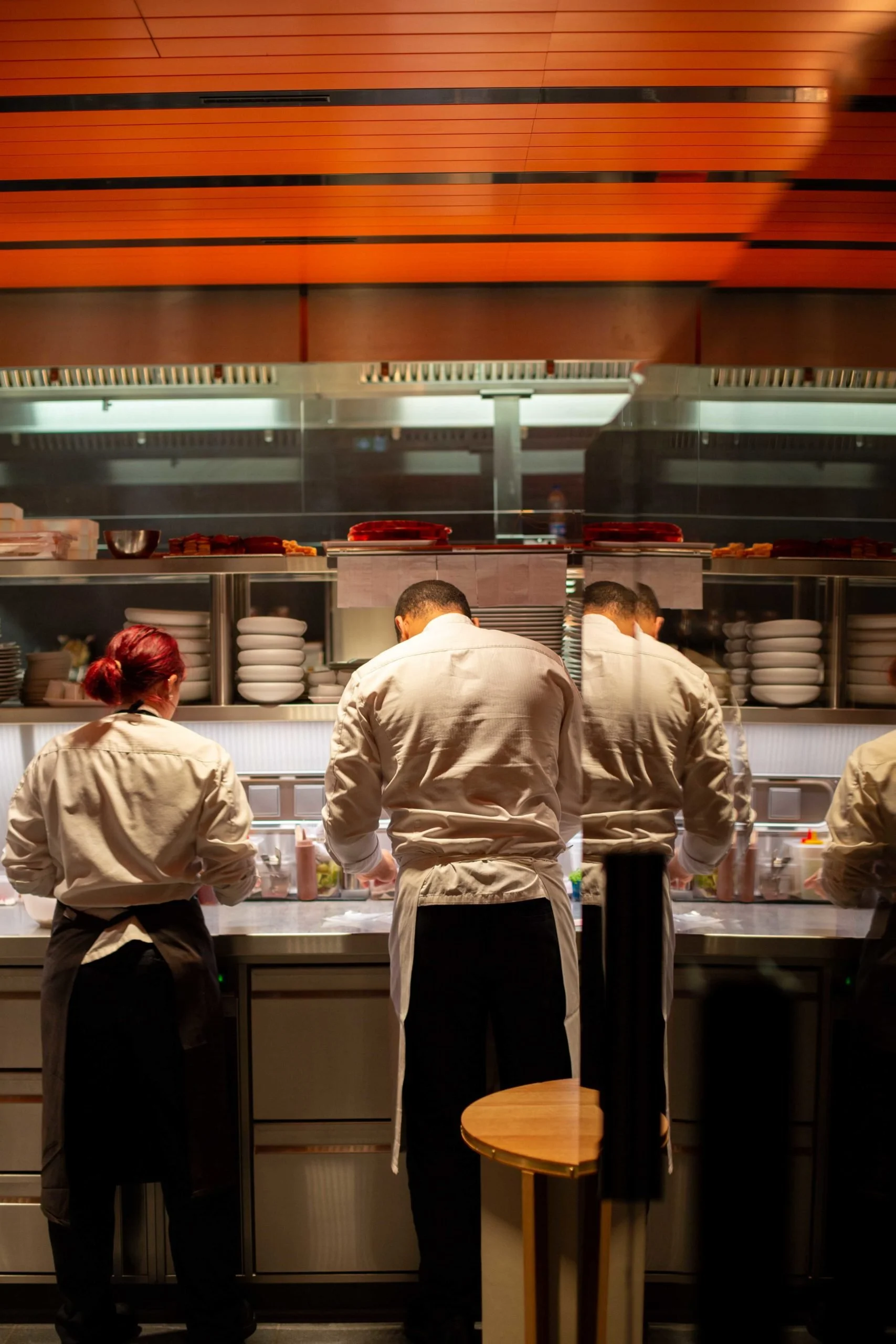 Trois chefs en tenue de cuisine travaillent au comptoir dans une cuisine de restaurant, avec un mur en bois en arrière-plan.