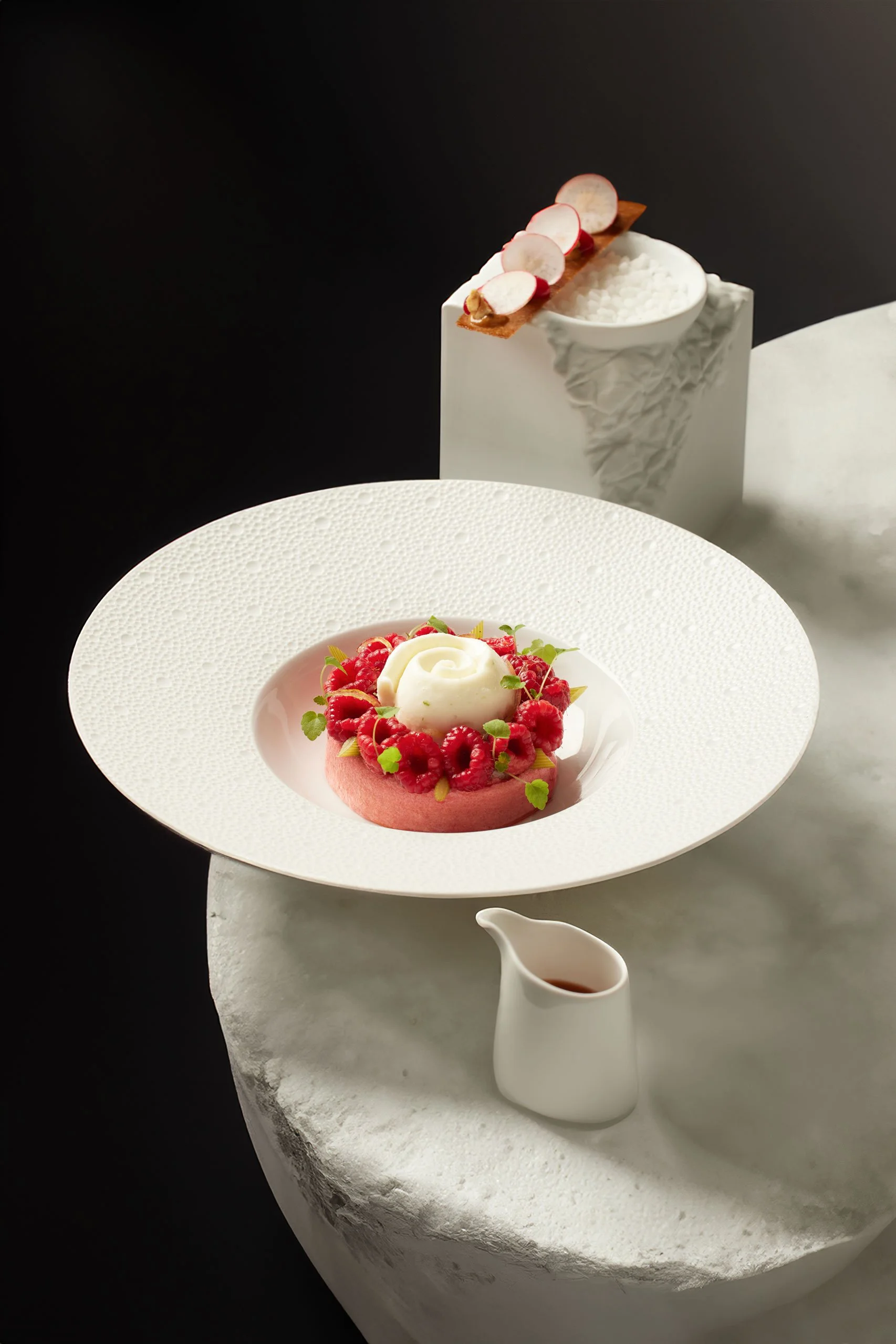 Une présentation élégante d'un dessert principal avec des framboises, une crème ou mousse blanche, et décorations de menthe, accompagnée d'un petit pot de sauce ou sirop, ainsi que deux autres desserts en arrière-plan, dont un avec des tranches de ra