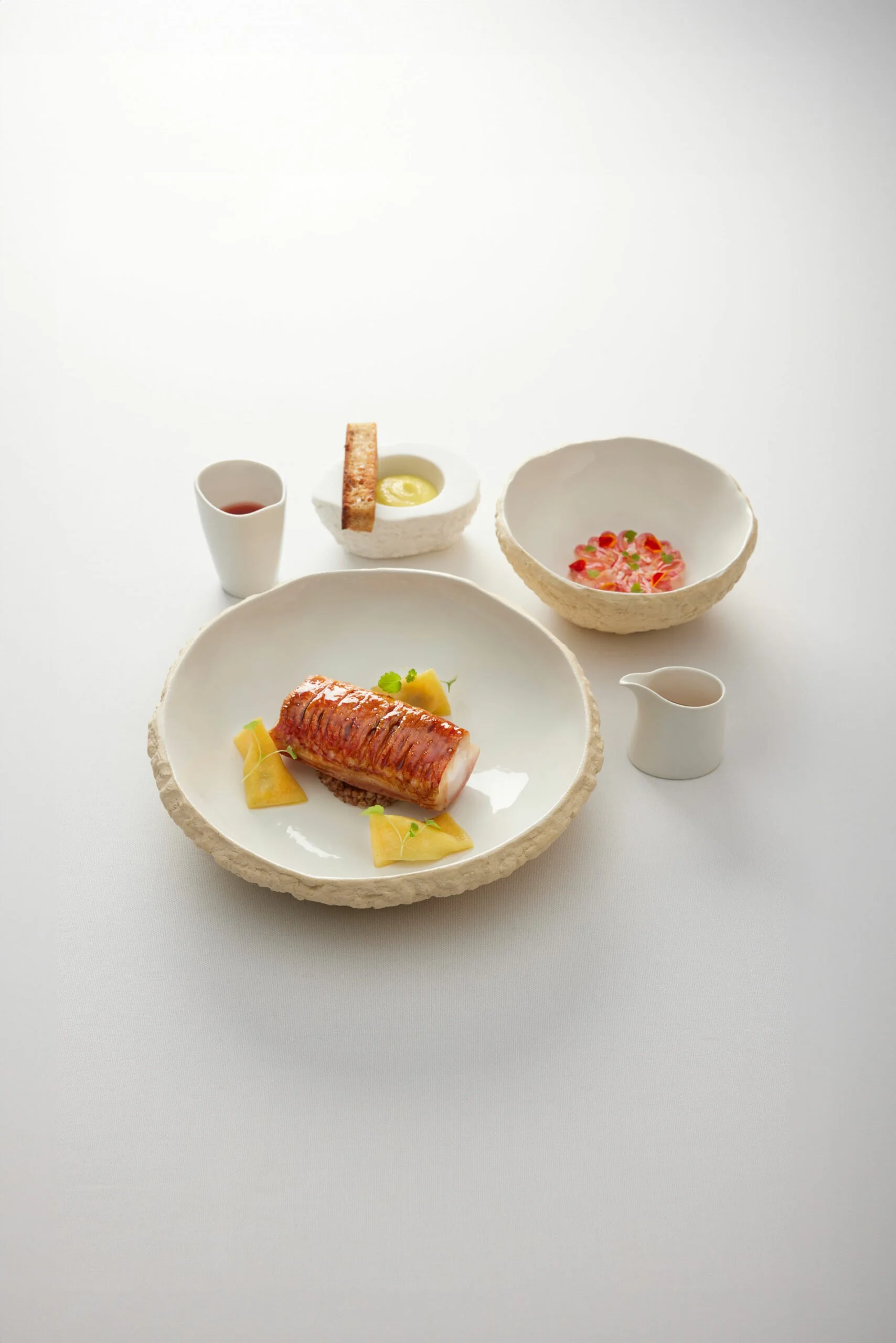 Un plat de cuisine gastronomique avec un morceau de poisson enroulé, servi avec des légumes et une sauce, dans une assiette blanche. Autour, des petits bols et saucières avec des accompagnements, sur une table blanche.