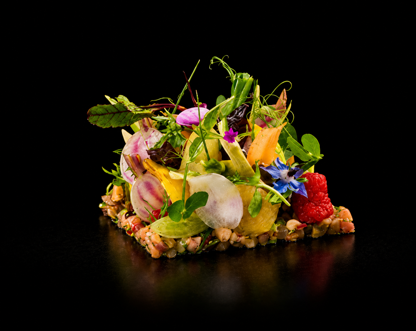 Salade délicate avec des légumes colorés, des herbes et des fleurs, présentée sur une assiette noire.
