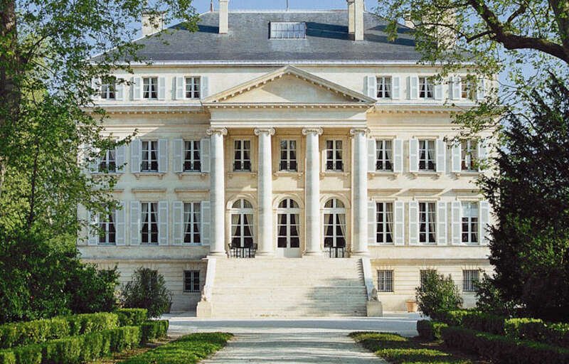 Vue frontale d'un grand bâtiment blanc avec colonnes et escaliers en pierre, entouré d'arbres et de jardins bien entretenus.