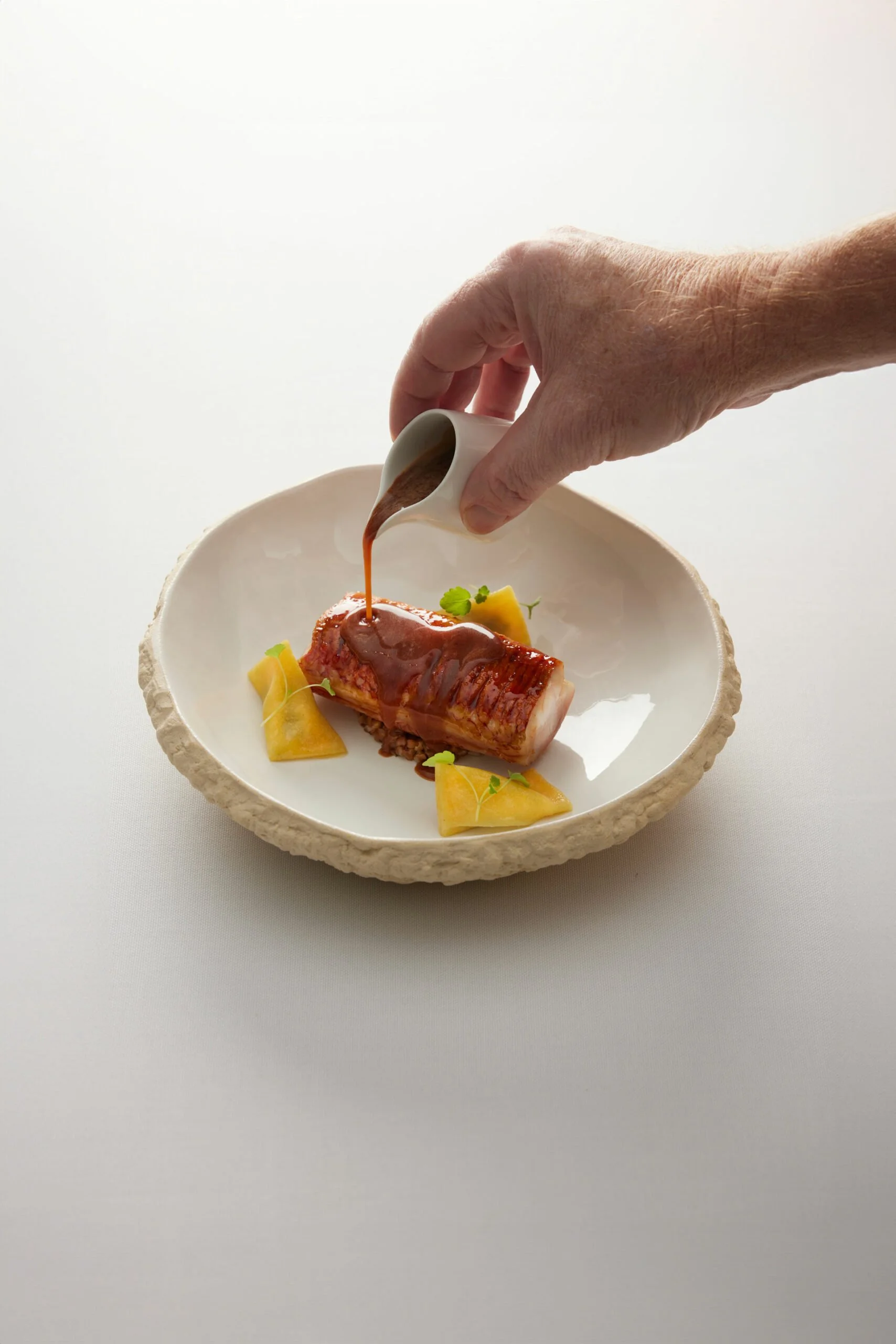 Une main verse une sauce brune sur un plat de viande enroulée, accompagné de petites portions de légumes jaunes, dans un bol blanc avec un rebord rugueux.