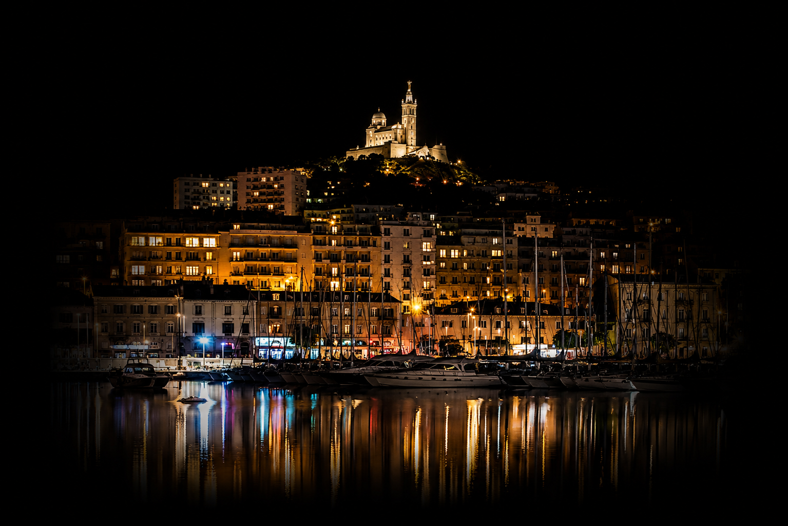 Vue nocturne d'un port avec des bateaux amarrés, bâtiments illuminés, et une basilique sur une colline éclairée en arrière-plan.