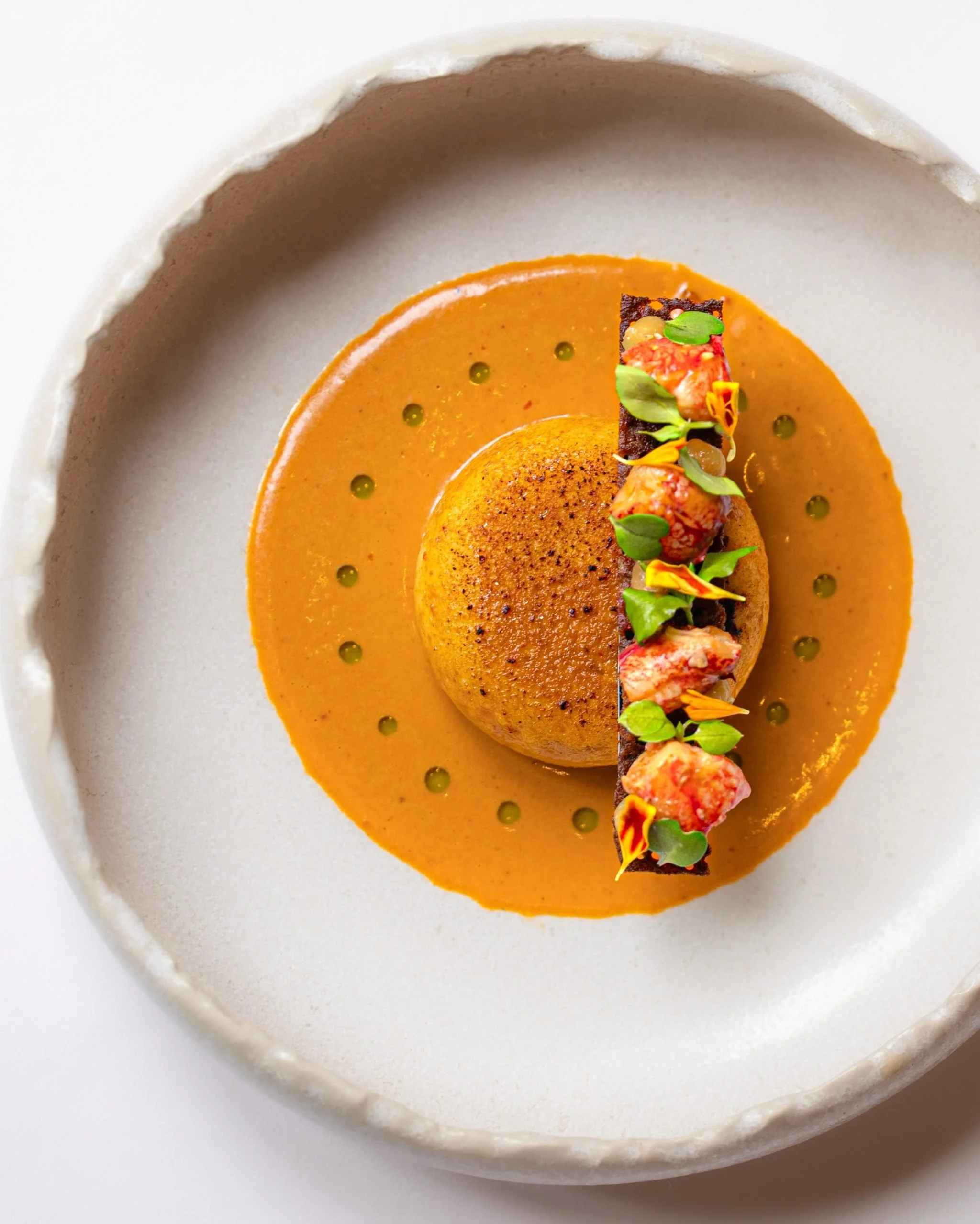 Plat gastronomique avec un rectangle de viande garnie de légumes colorés et de fleurs comestibles, entouré de sauce orange.