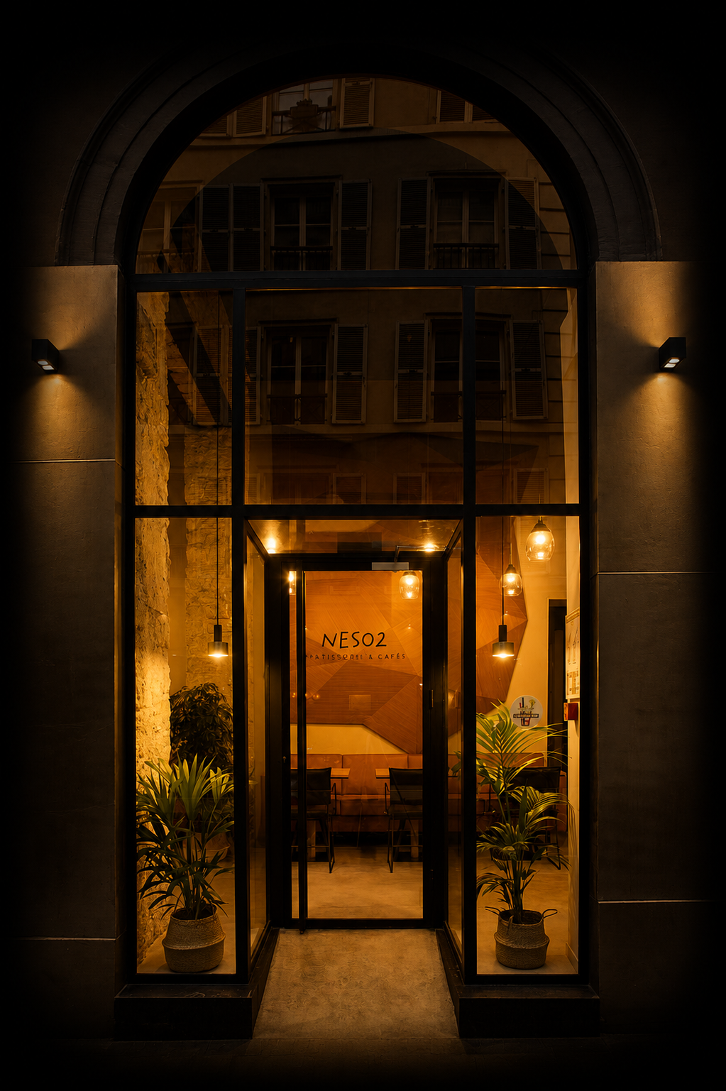 Vue de la façade d'une boutique ou café appelée NESO2, avec une grande porte en verre encadrée en noir, deux plantes en pot de chaque côté, et un éclairage chaleureux à l'intérieur.