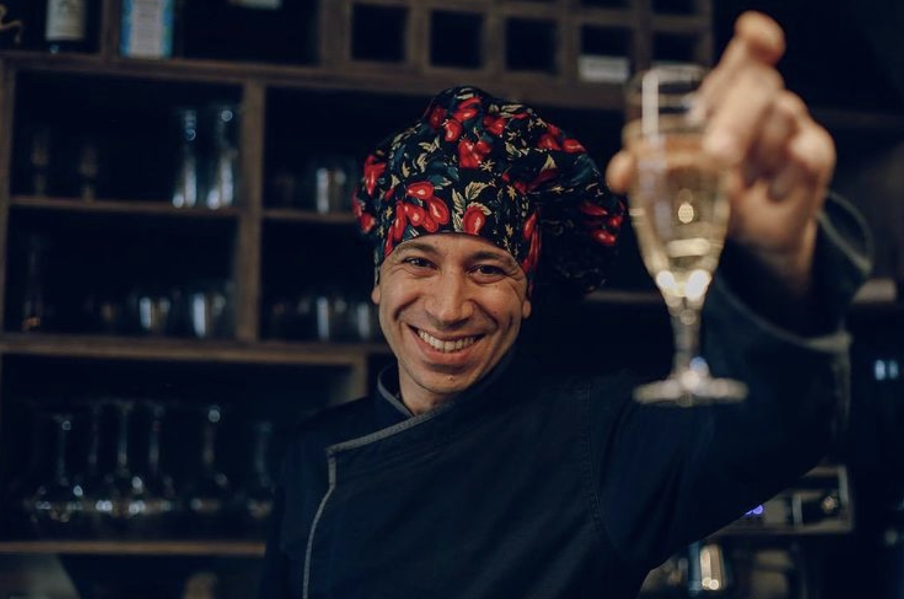 Un chef souriant portant un toque à motifs floraux fait un toast avec un verre à vin dans une cuisine ou un bar.