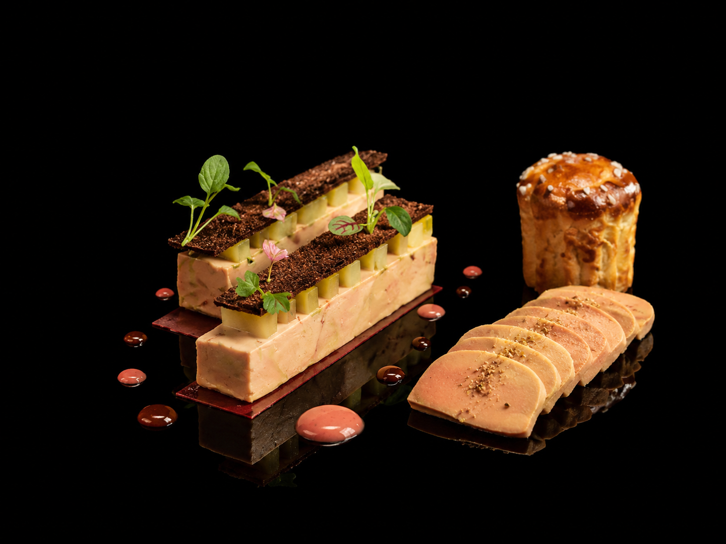 Assortiment de pâtisseries décoratives sur fond noir, comprenant une terrine de foie gras, un pain d'épices, et des tranches de pâté, avec des décorations de microgreens et de petites sauces.