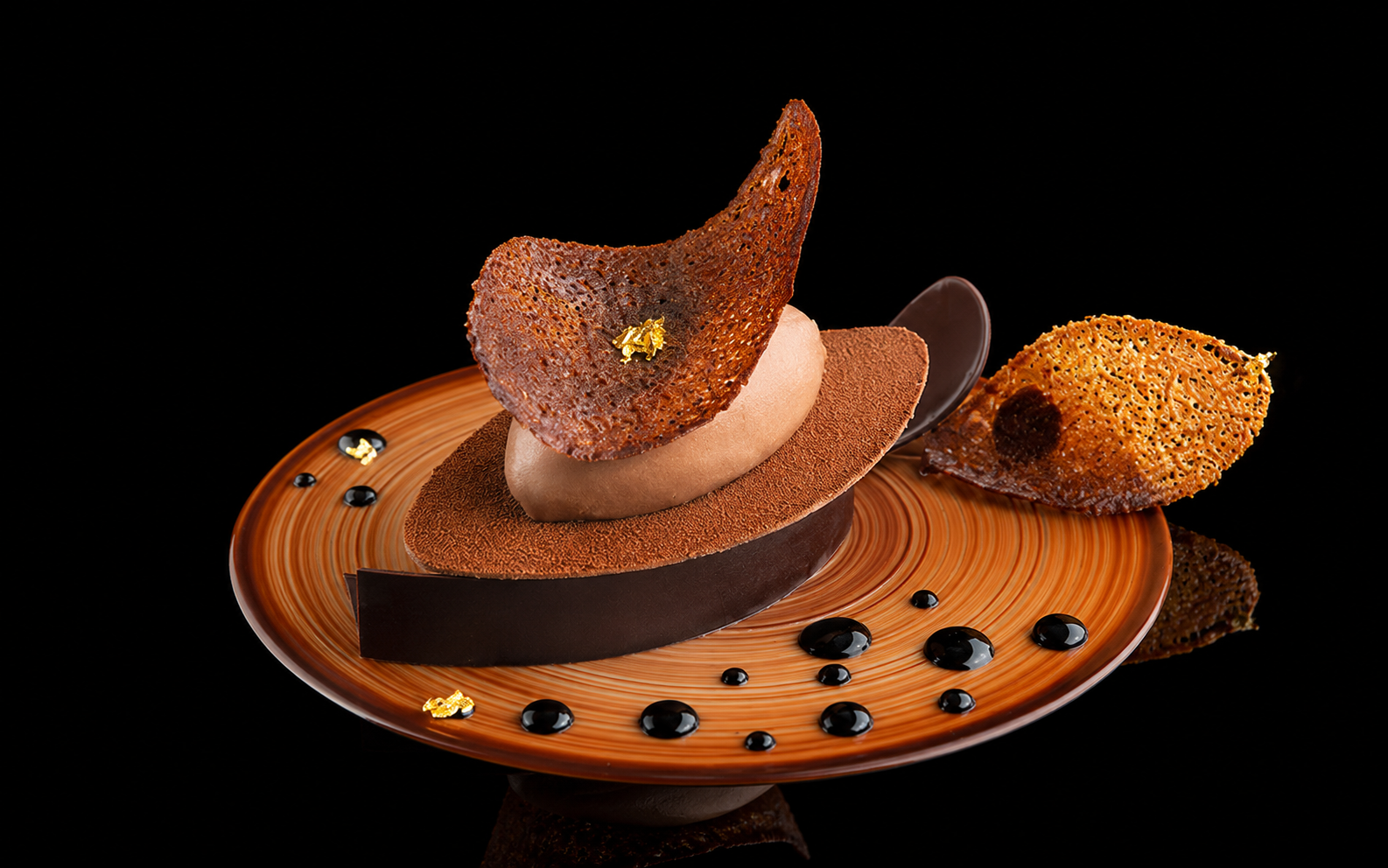 Un dessert sophistiqué avec une mousse au chocolat, une fine feuille de chocolat noir, une décoration en caramel croustillant et des gouttes de sauce chocolat sur une assiette orange avec un fond noir.