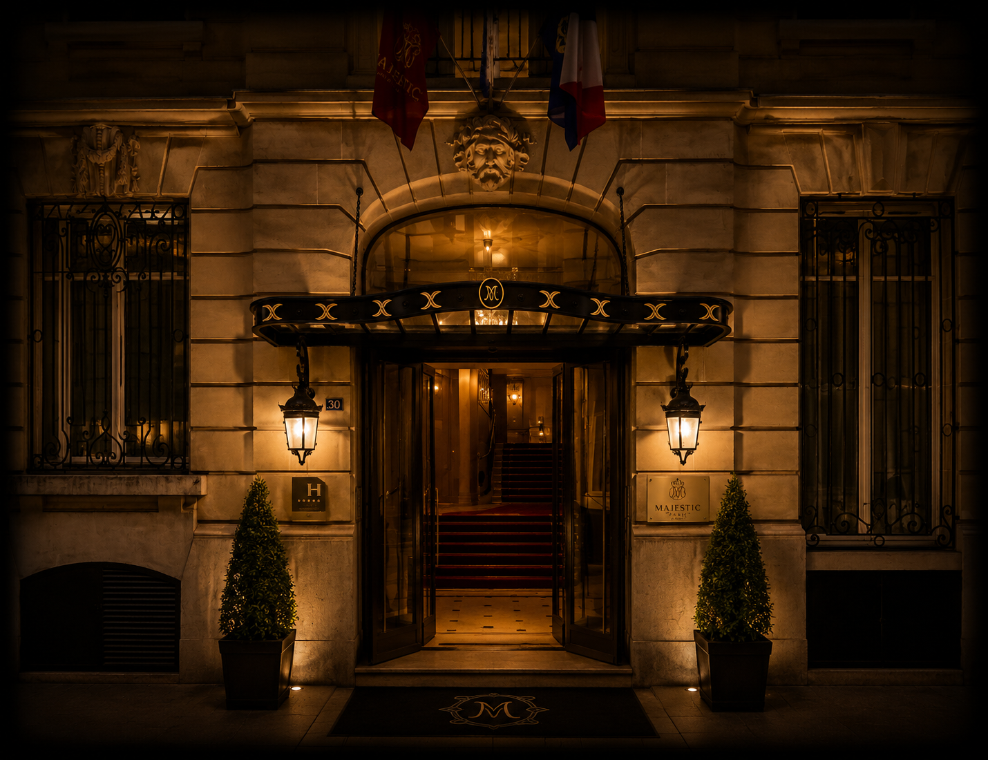 Entrée d'un hôtel de luxe, façon Paris, à la nuit. Luminosité chaude, deux lampes extérieures, deux pots de buissons, portes en verre ouvertes, escalier intérieur enroulé, drapeaux français et autres, sculptures et décorations classiques.