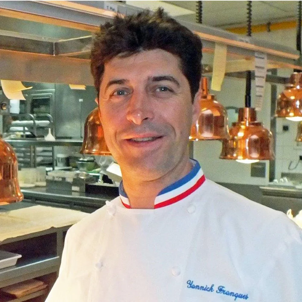 Chef professionnel portant une veste blanche avec le nom Hannick Franques, dans une cuisine équipée et moderne.