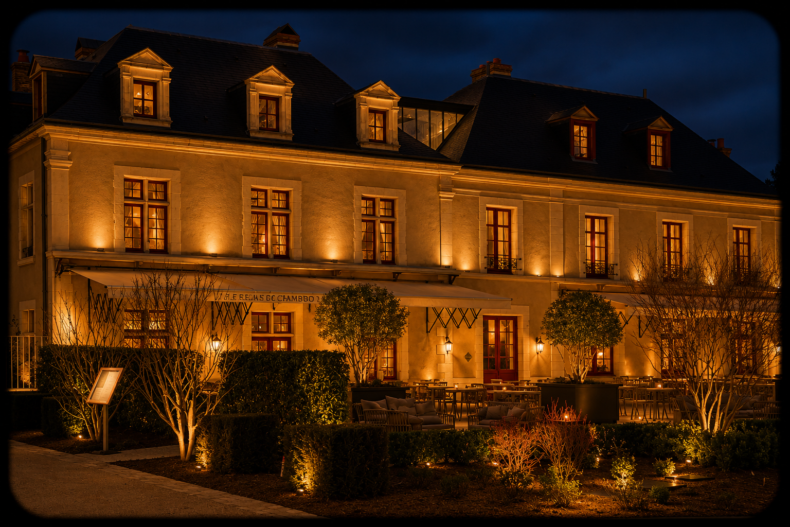 Un bâtiment élégant éclairé la nuit, avec des fenêtres illuminées et un espace terrasse avec des tables, des chaises, des arbres arrosés par des lumières extérieures, dans un style parisien classique.