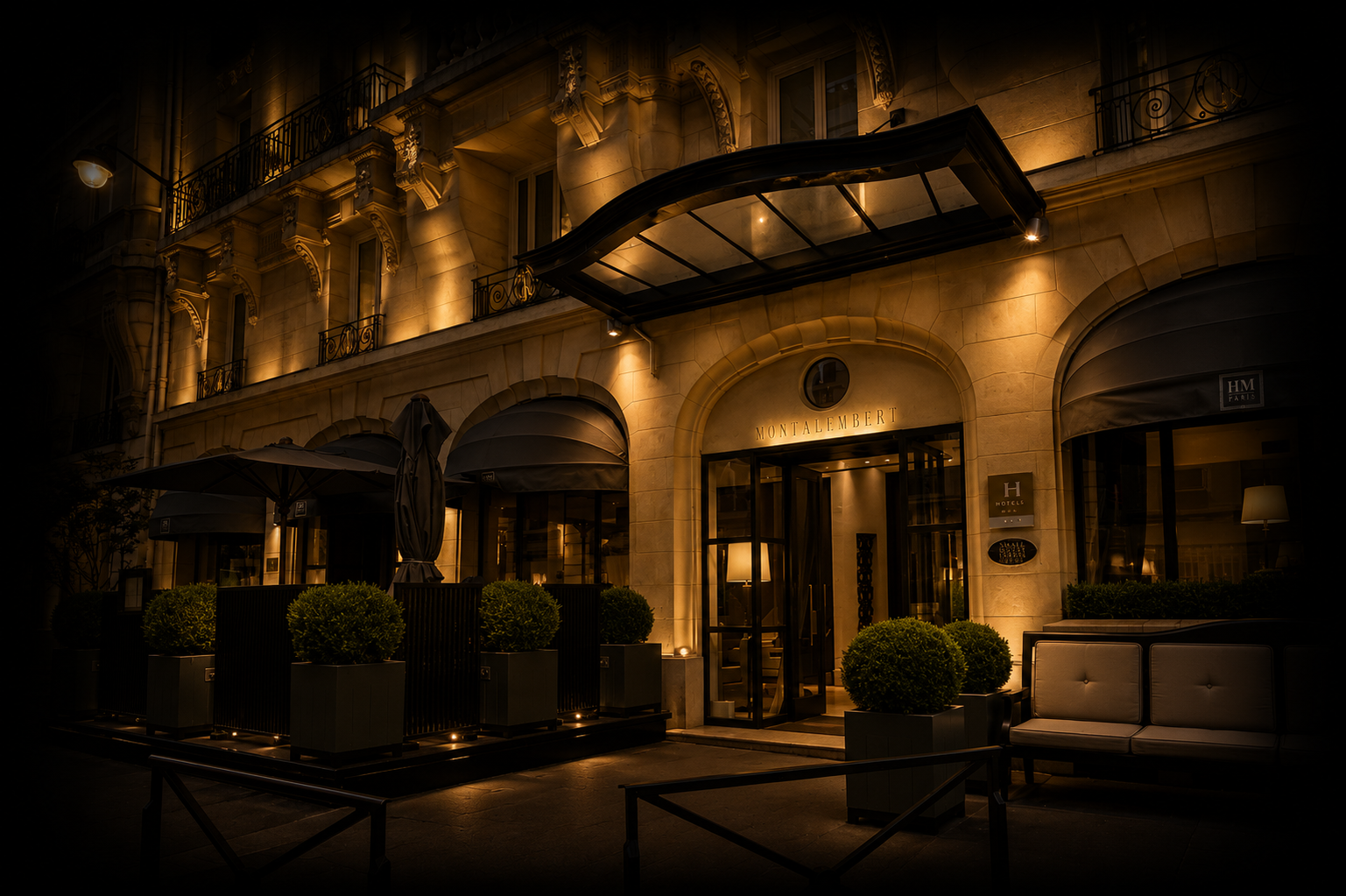 Façade d'un hôtel élégant et lumineux la nuit, avec une enseigne 'Montalembert' au centre, des plantes en pots, des fauteuils, et des fenêtres avec des balustrades en fer forgé.