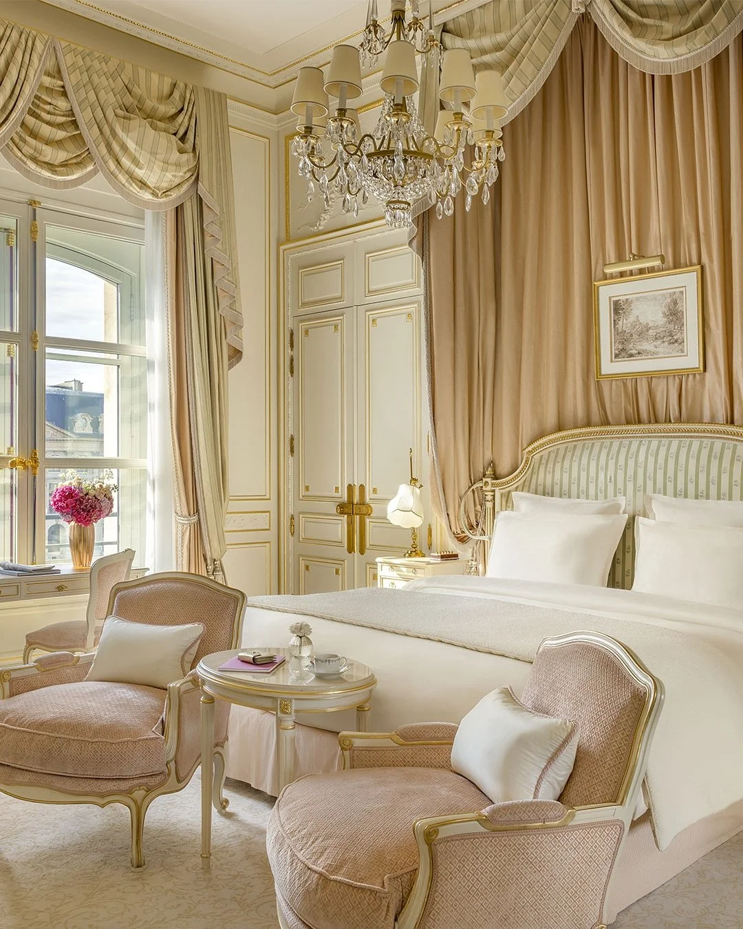 Chambre élégante avec un lit à baldaquin, fauteuils en tissu rose, table ronde, lampadaire, rideaux en tissus crème et beige, lustre en cristal, et une fenêtre avec volets donnant sur l'extérieur.