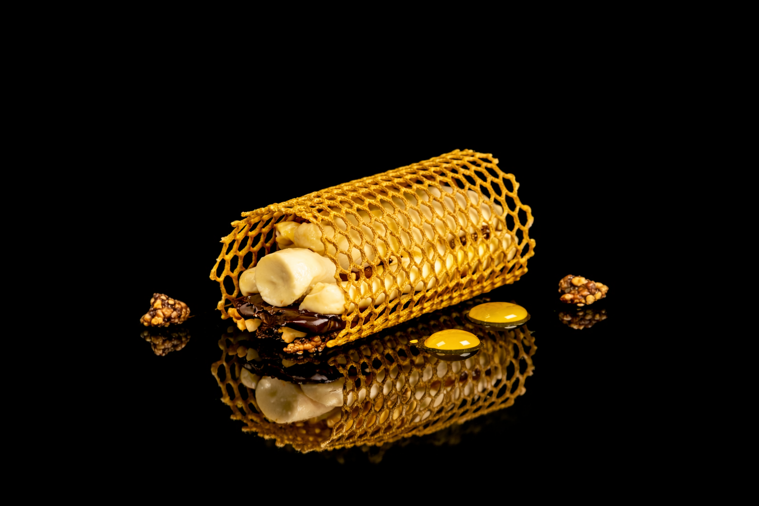 Désert sophistiqué avec un cornet croustillant rempli de crème, de chocolat et de noix, avec des éclats de noix, des cuillères de sauce jaune et des morceaux de chocolat, posé sur une surface noire miroir.