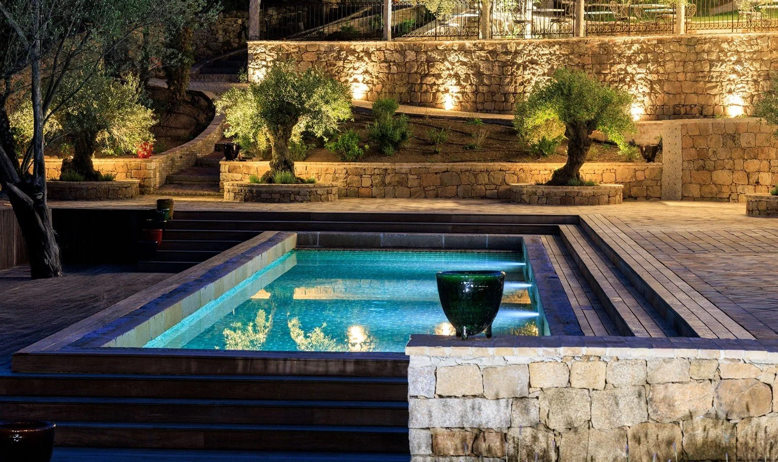 Piscine éclairée dans un jardin avec des arbres et un mureti en pierre, éclairés par des lampes.