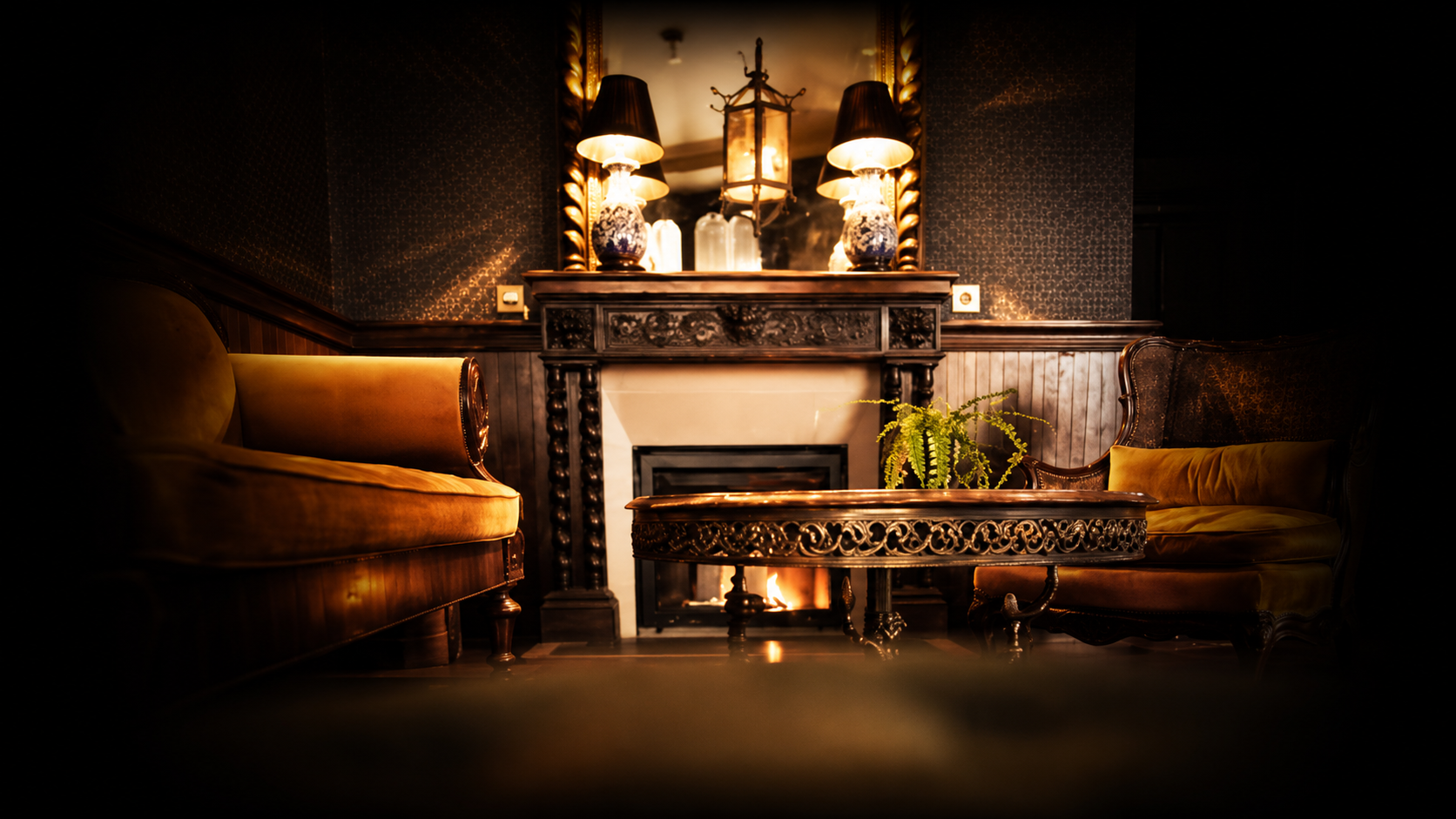 Intérieur chaleureux avec un cheminée en marbre noir et bois sculpté, deux sofas en velours doré, une table en bois finement sculptée, une plante verte, un grand miroir encadré en or, deux lampes en porcelaine bleue et blanche, une suspension en fer avec des vitrines derrière.