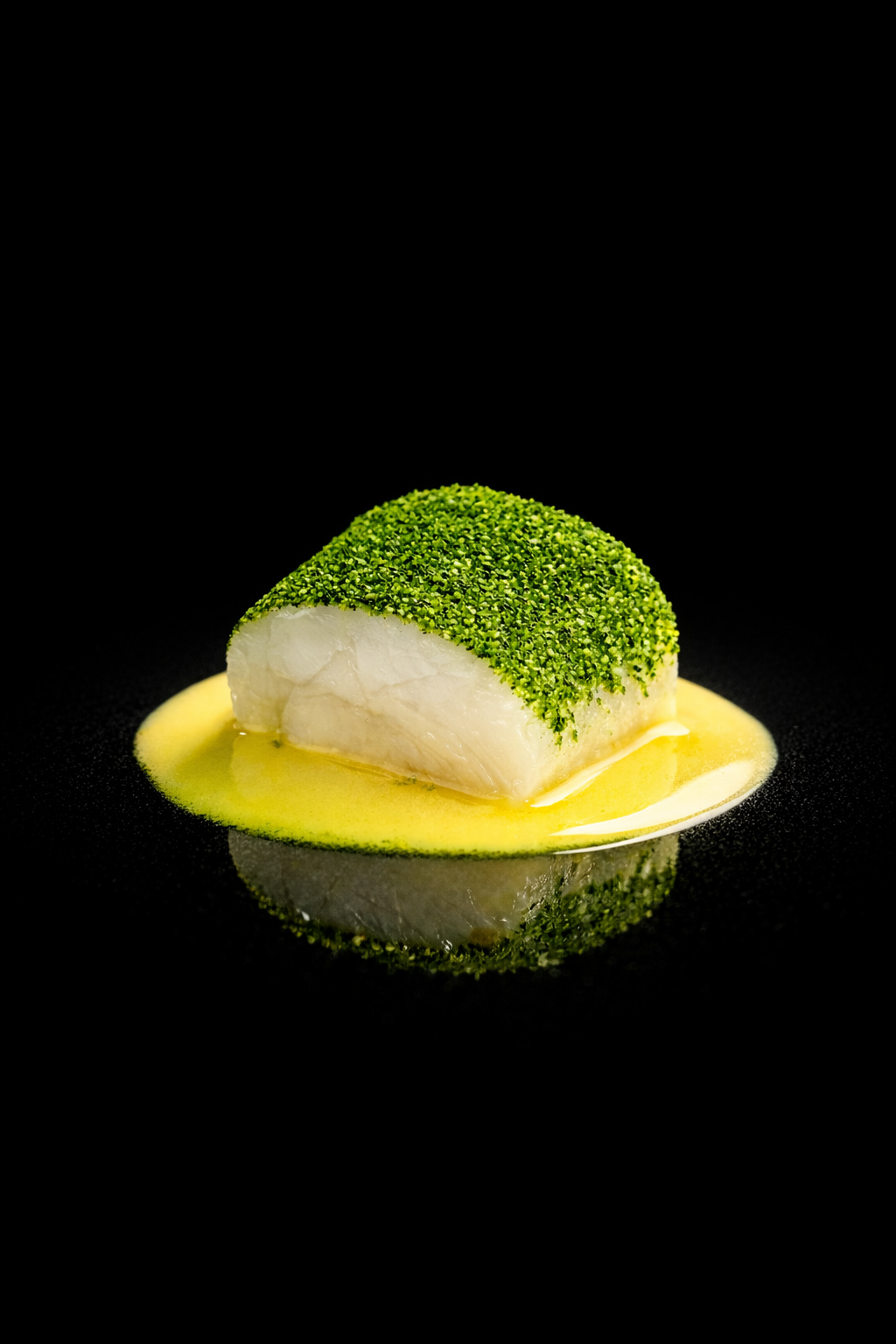 Poisson blanc sur une assiette noire, recouvert de pistaches, avec une sauce jaune.