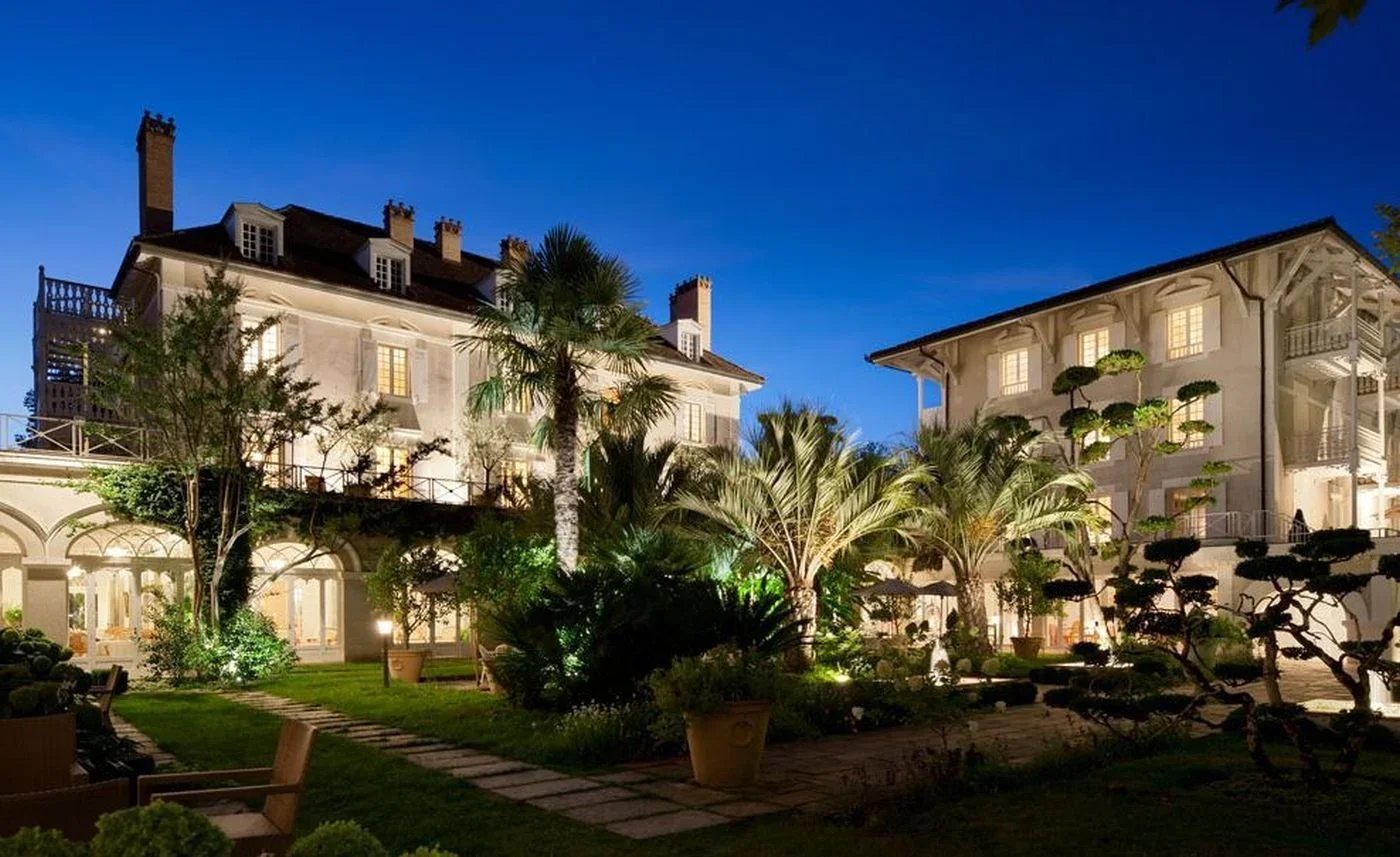 Maisons de style européen avec jardin éclairé la nuit, comprenant des palmiers et arbres taillés, sous un ciel nocturne.