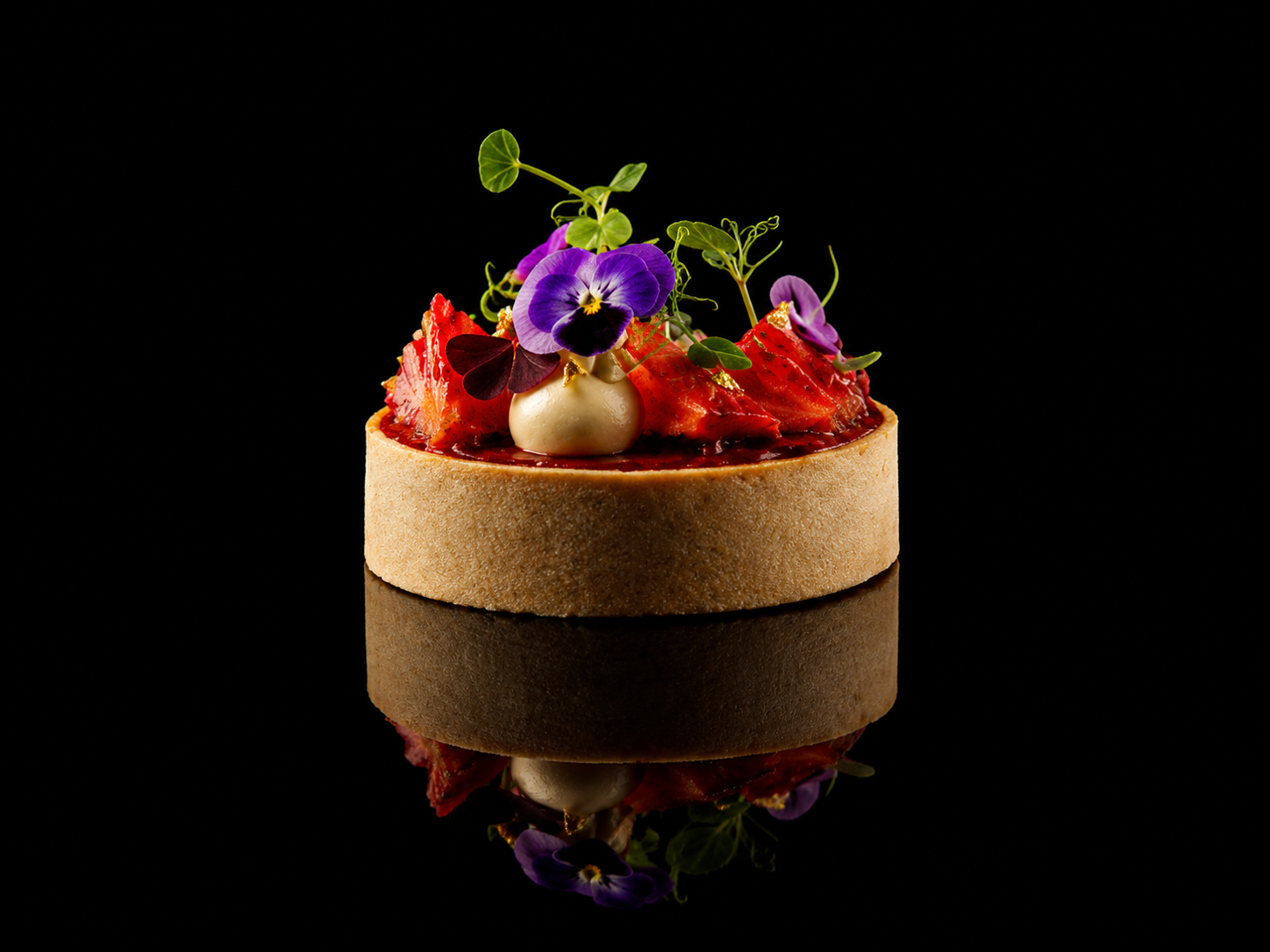 Gâteau sophistiqué décoré de fraises tranchées, fleurs violettes et vertes, avec une base circulaire en biscuit, sur fond noir avec reflet au sol.