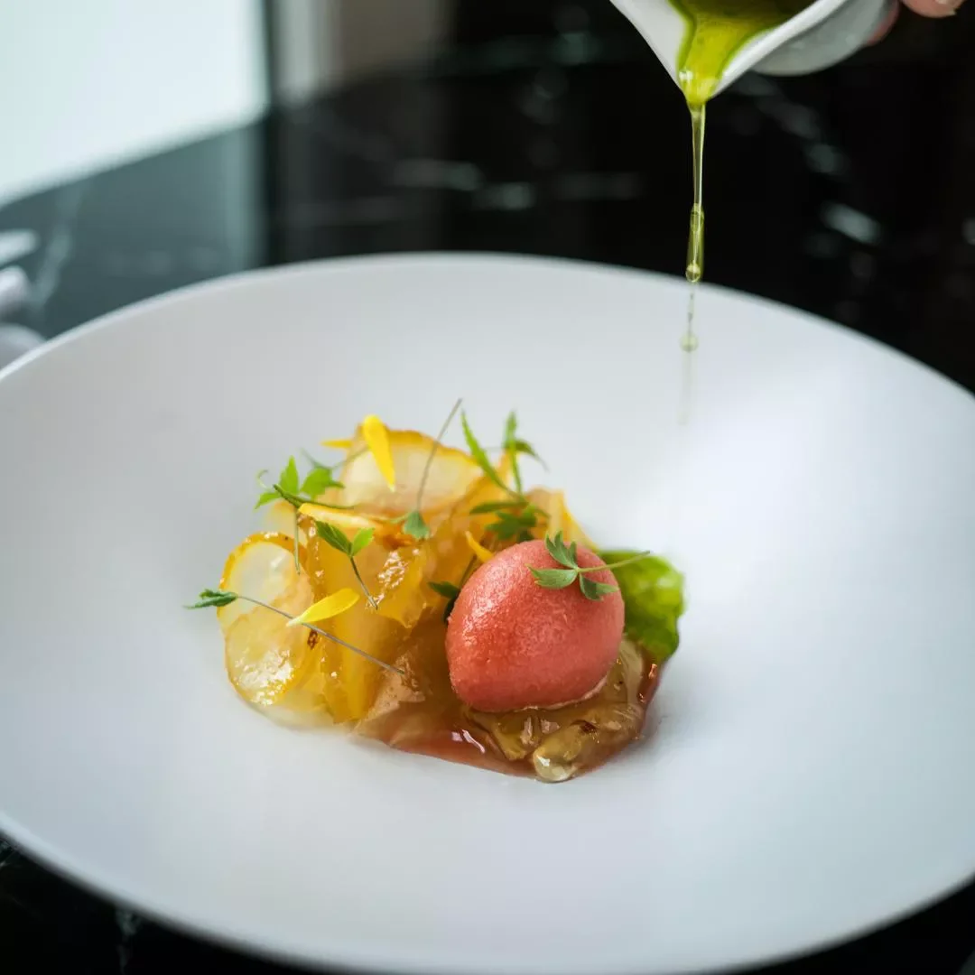 Un plat gourmet avec une boule de glace ou sorbet rose, garniture de zestes de citron, feuilles vertes, et une sauce ou gelée translucide, avec de l'huile d'olive versée au-dessus.