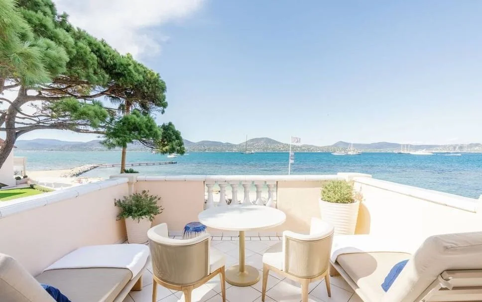 Terrasse en bord de mer avec vue sur la mer et îles, mobilier de patio blanc, chaises et table, plantes en pot, ciel clair.