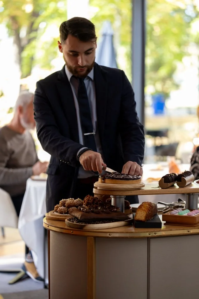 Un homme en costume coupe un gâteau dans un café ou restaurant avec des desserts et cookies sur une table en bois.