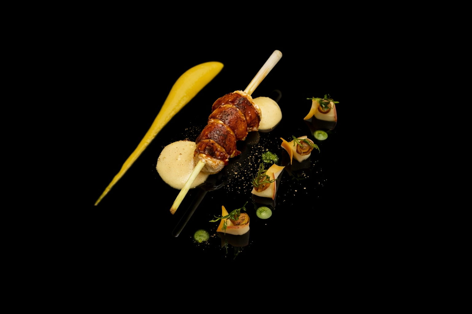 Assiette gastronomique avec une brochette de viande, purée, sauce verte, et petites décorations végétales sur un fond noir.