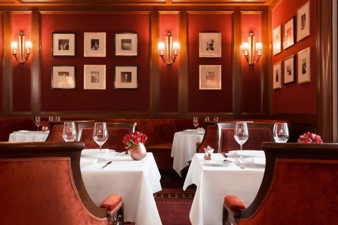Salle à manger élégante avec murs rouges, tableaux en noir et blanc, chandeliers et tables avec nappes blanches, verres à vin et fleurs rouges.