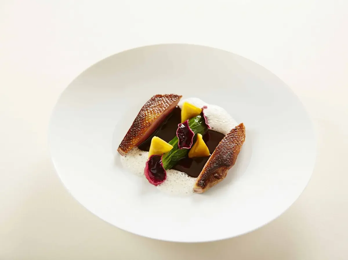 Assiette de dessert avec deux morceaux de mangue, une feuille de menthe, une sauce au chocolat, une crème mousseuse et des fleurs comestibles