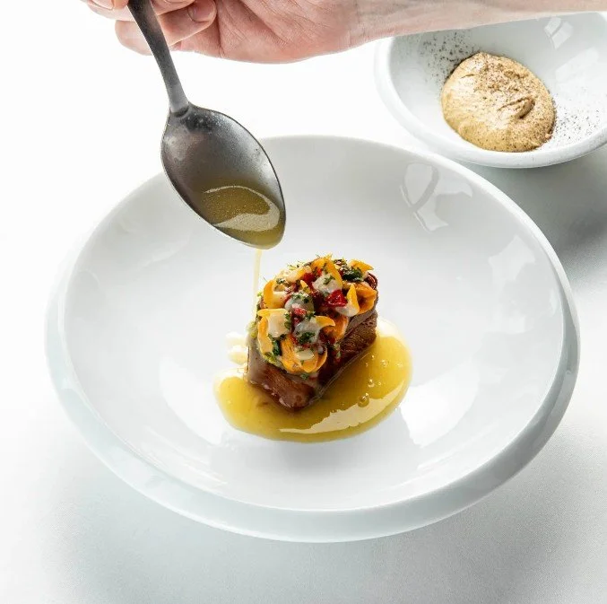 Une main verse une sauce sur un plat de viande avec des légumes, dans une assiette blanche. En arrière-plan, un bol contenant une épice en poudre.