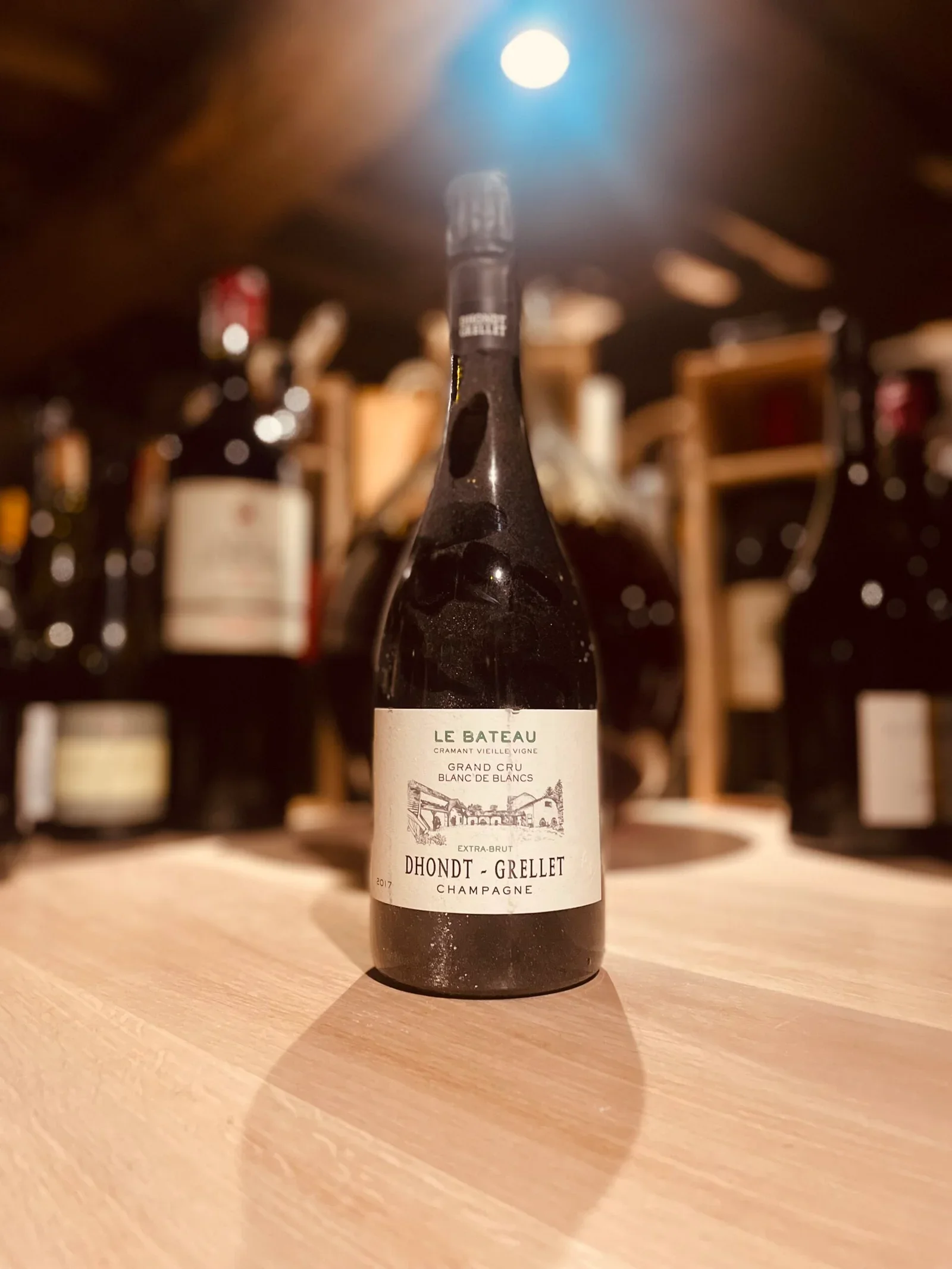 Bouteille de champagne Dhondt-Grellet sur une table en bois, fond flou avec plusieurs autres bouteilles de vin dans une cave ou un bar.