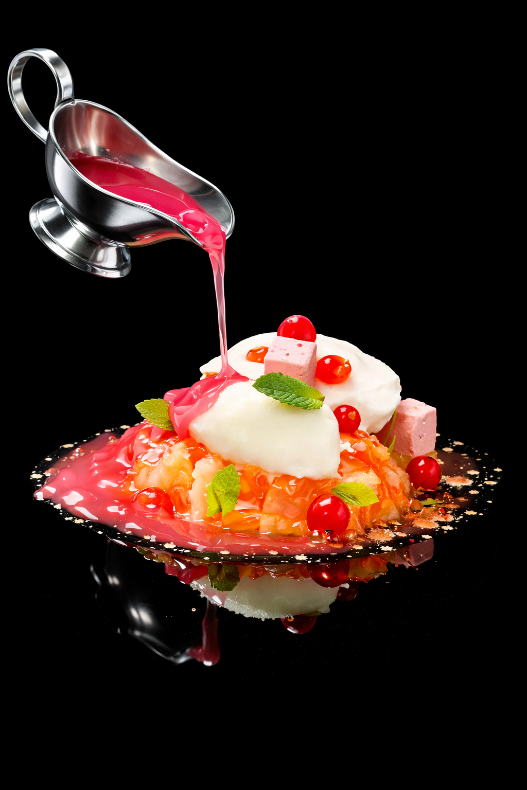 Un dessert moderne avec une sauce rouge being versée dessus, décoré de morceaux de fruits, de cubes de pâtisserie, de feuilles de menthe et de petits fruits rouges, présenté sur une assiette noire reflective.