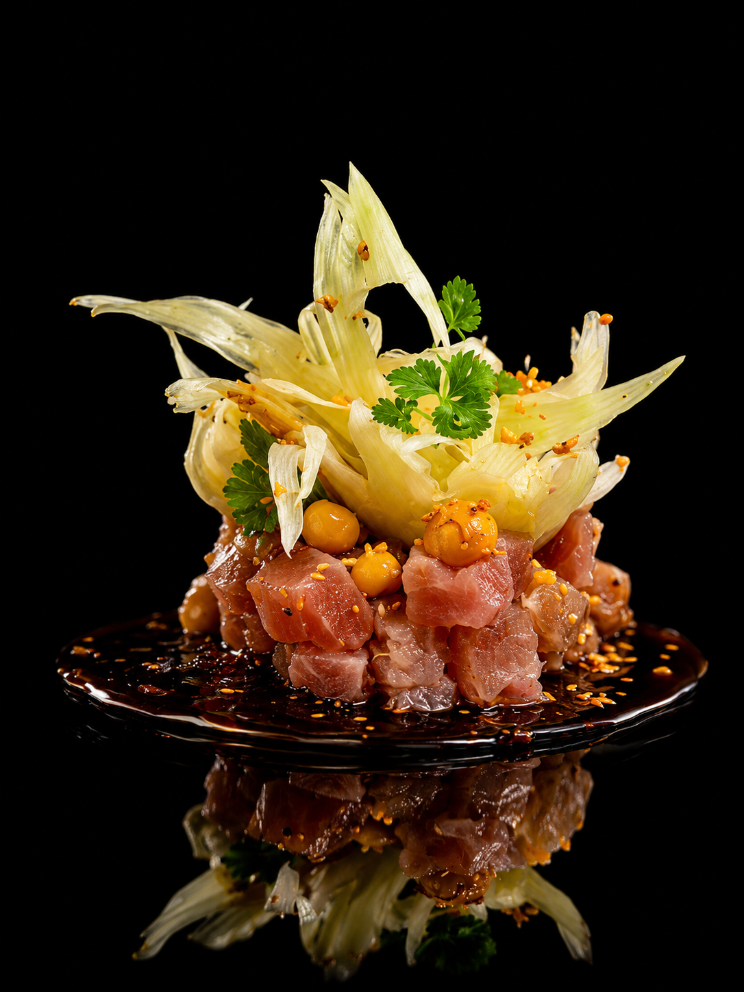 Un plat de tartare de poisson avec du gingembre, cilantro, et une décoration de fenouil sur une assiette en verre noir, reflet visible en dessous.
