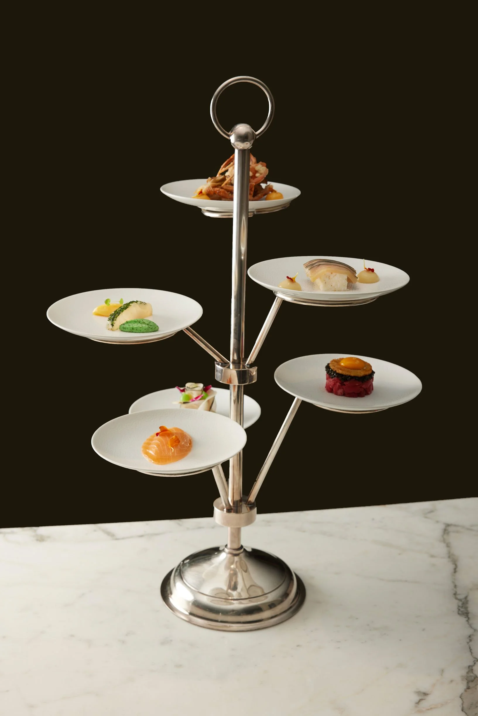 Plateau de dégustation avec plusieurs petits plats raffinés sur une structure à plusieurs niveaux en métal, posé sur une table en marbre blanc, fond noir.
