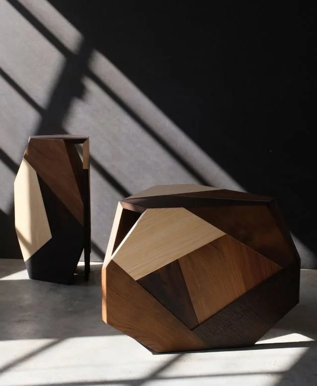 Deux tables en bois géométriques. L'une est debout, l'autre est inclinée sur le sol, avec des ombres de fenêtres projetées sur le mur et le sol.