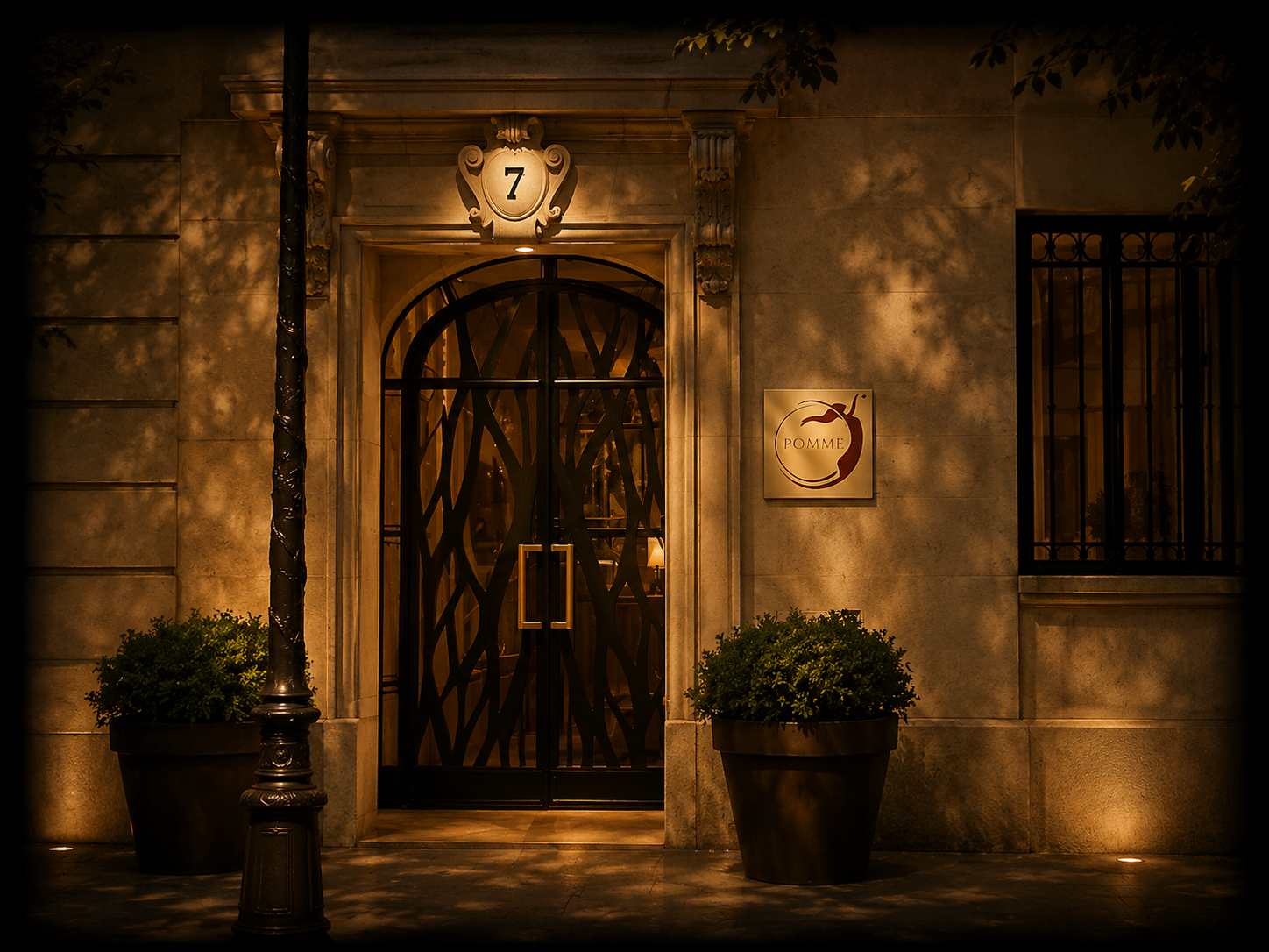 Façade d'une boutique ou restaurant appelé Pomme, avec une porte en fer décoratif, une enseigne avec une pomme stylisée et des pots de fleurs en façade, éclairée chaleureusement la nuit.
