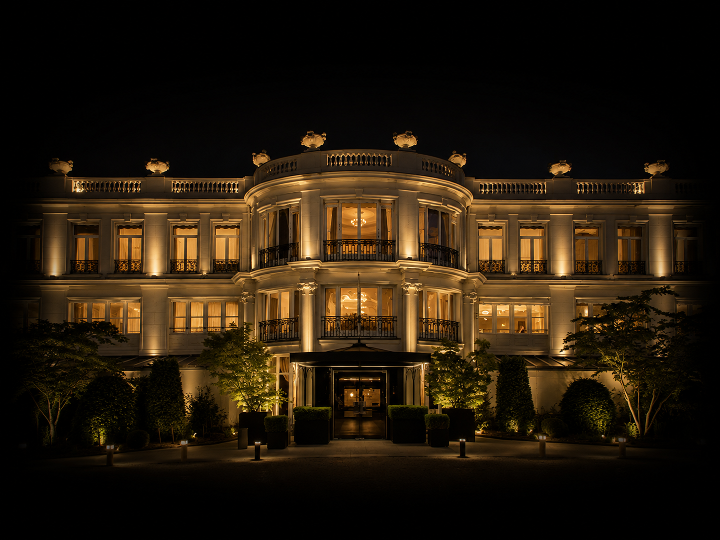 Vue nocturne d'un grand hôtel élégant illuminé avec des fenêtres éclairées, un jardin soigné avec des arbres et des lumières extérieures.
