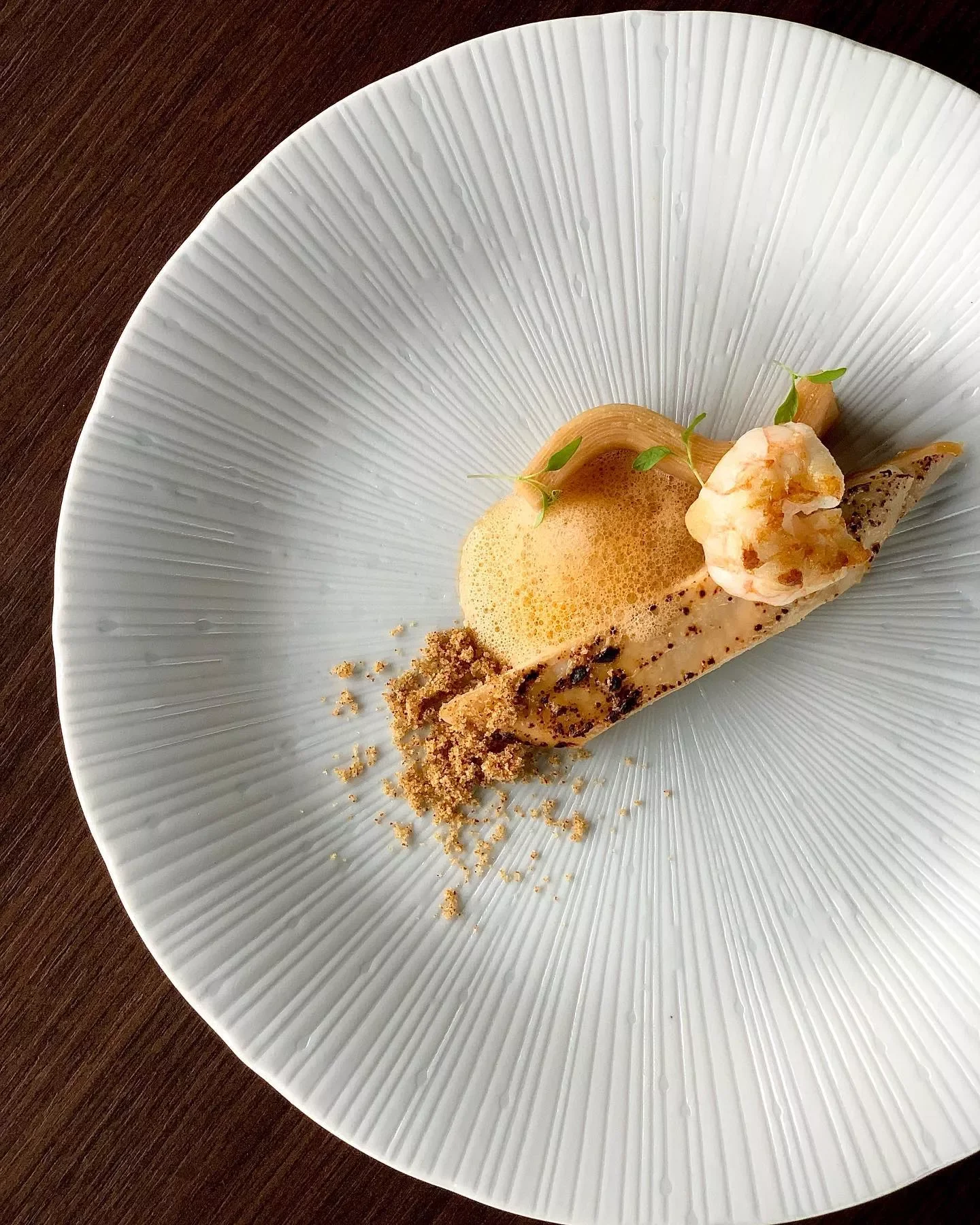 Un plat gastronomique présenté artistiquement avec une sauce mousseuse, une crevette, et des garnitures vertes sur une assiette blanche à rayures légères.