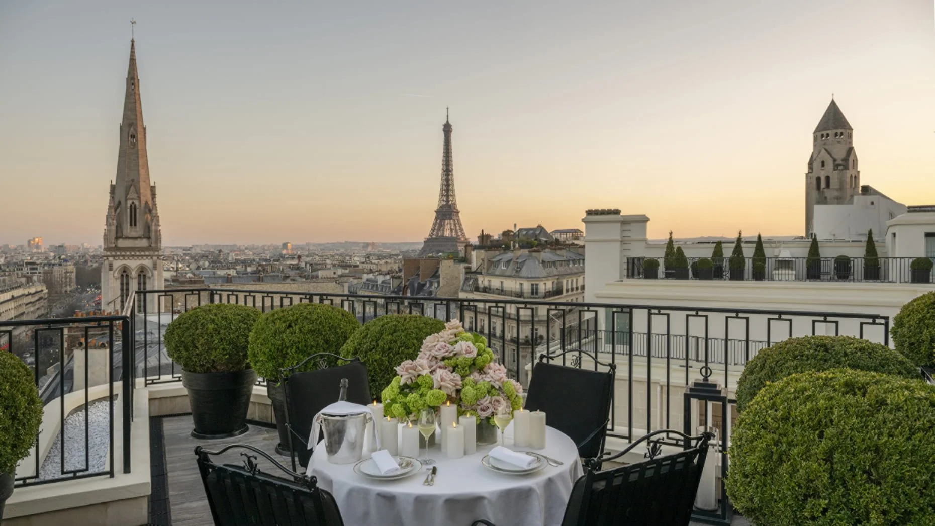 Vue d'une terrasse parisienne avec une table dressée pour un repas, entourée de plantes en pot, avec la tour Eiffel et des églises en arrière-plan au coucher du soleil.