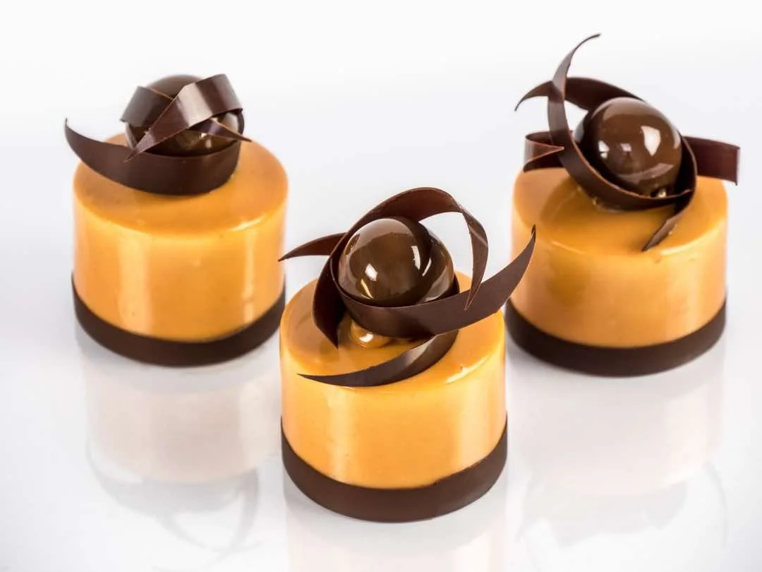 Trois desserts modernes avec une base en mousse orange, décorés de sphères en chocolat et de spirales en chocolat