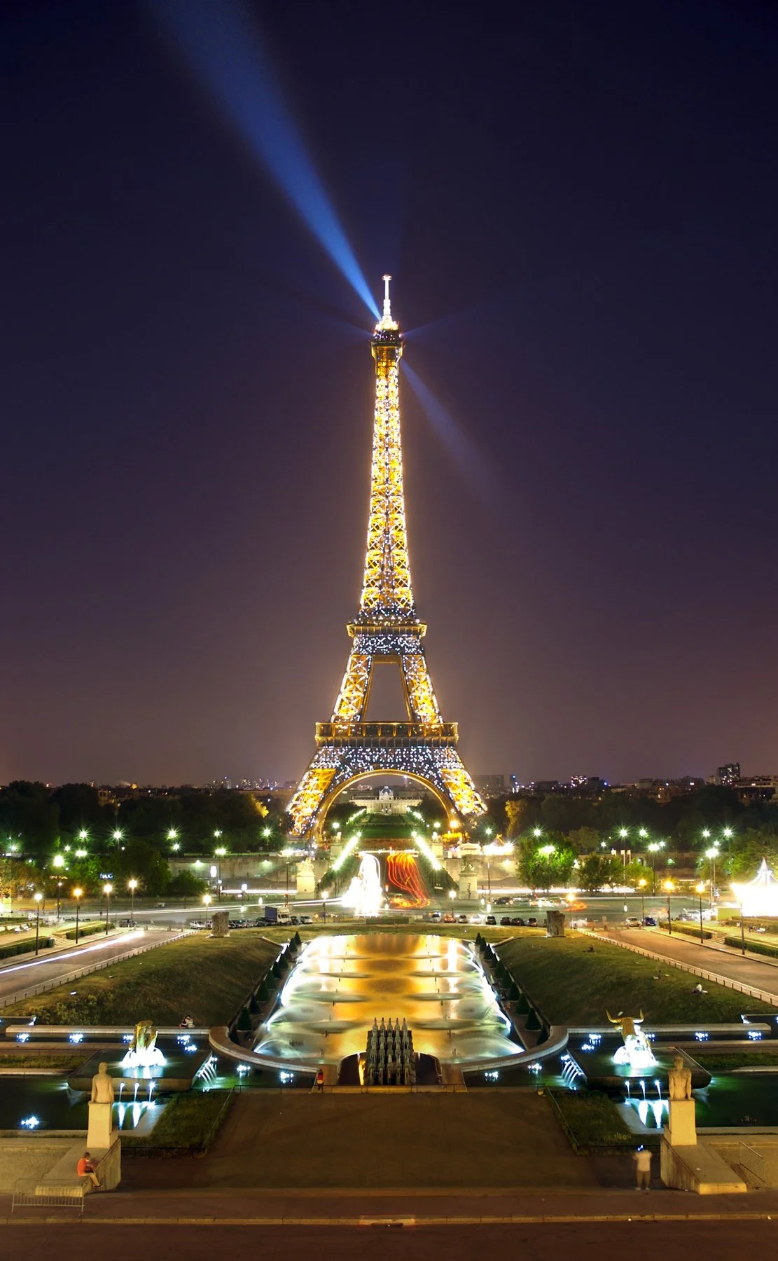 Tour Eiffel illuminée la nuit avec un éclairage doré, reflet dans une fontaine au premier plan, ville de Paris en arrière-plan.