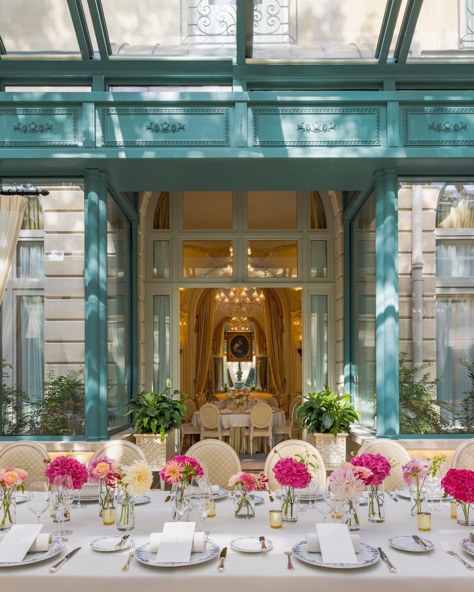 Salle de réception élégante avec table décorée de fleurs roses et blanches, vue à travers une verrière d'un intérieur somptueux avec chandelier chandelier chandelier et décoration luxueuse.