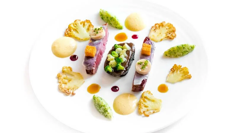 Assiette avec des petits morceaux de viande, des légumes en purée, et des décorations de légumes, sauce et crème en garniture.