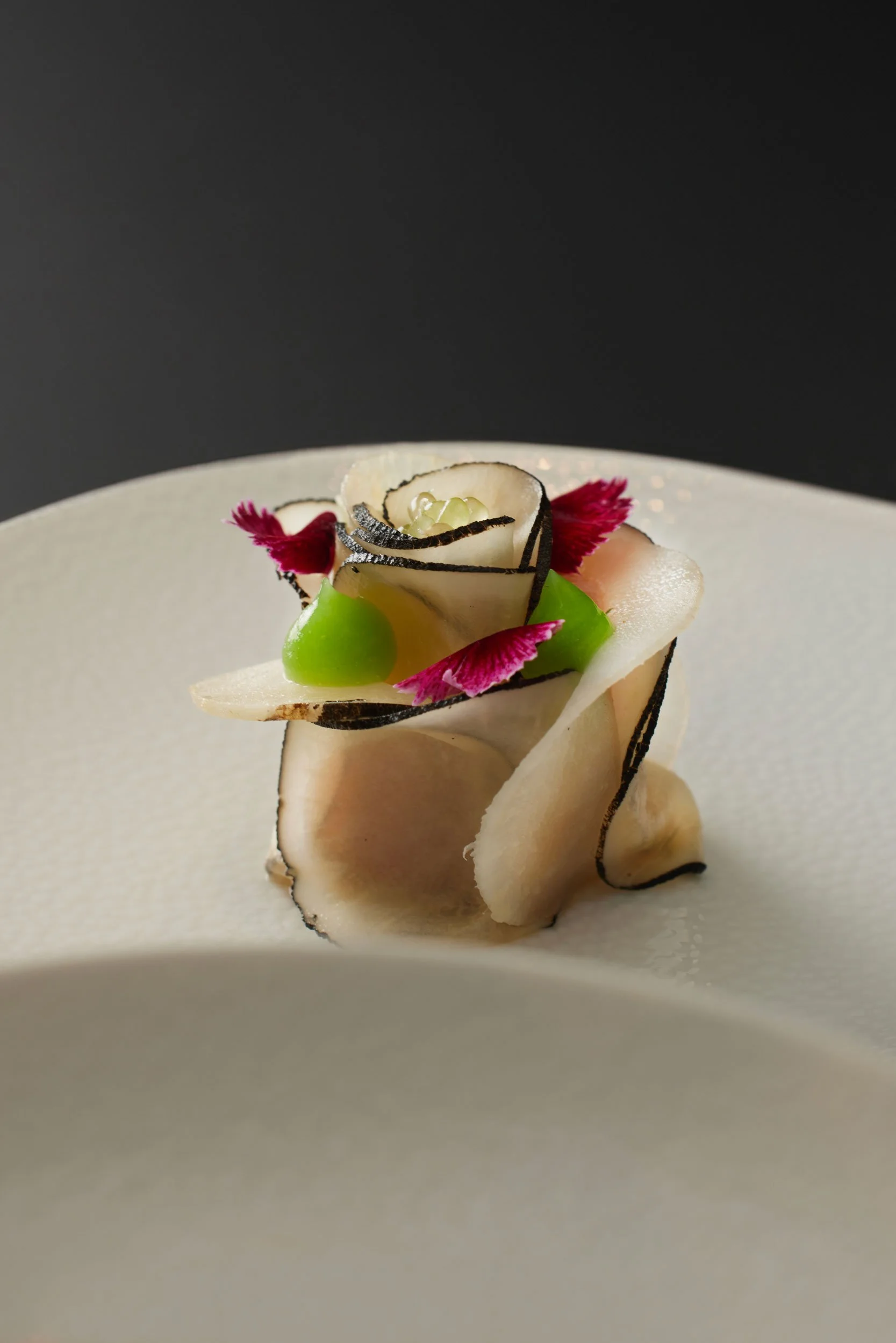 Un plat de cuisine sophistiquée avec des fines tranches de légumes ou de champignons, décoré avec des petits éléments colorés et des pétales roses, présenté sur une assiette blanche.
